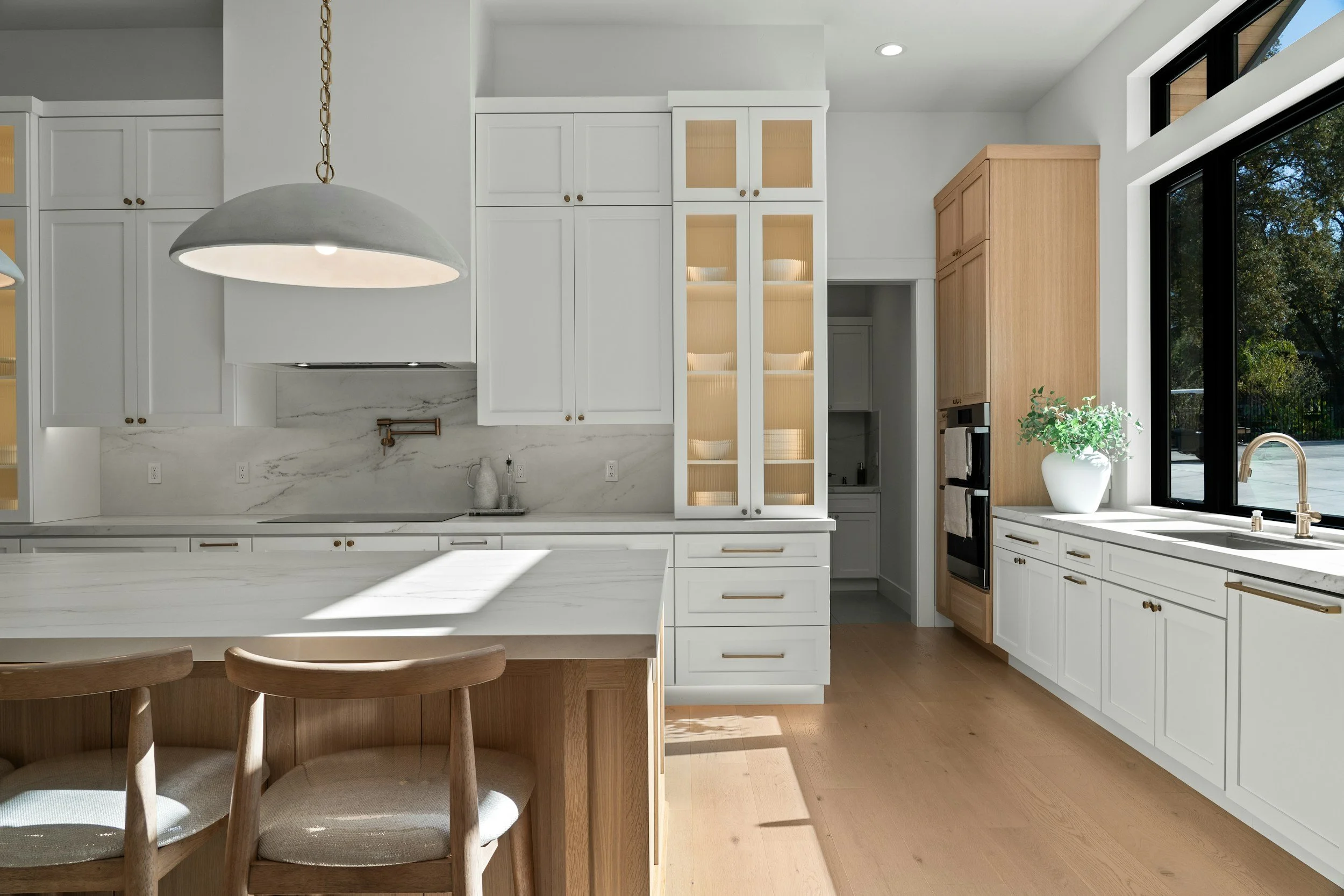 Modern kitchen with white cabinets, wooden accents, large window with trees outside, and a kitchen island with wooden chairs.