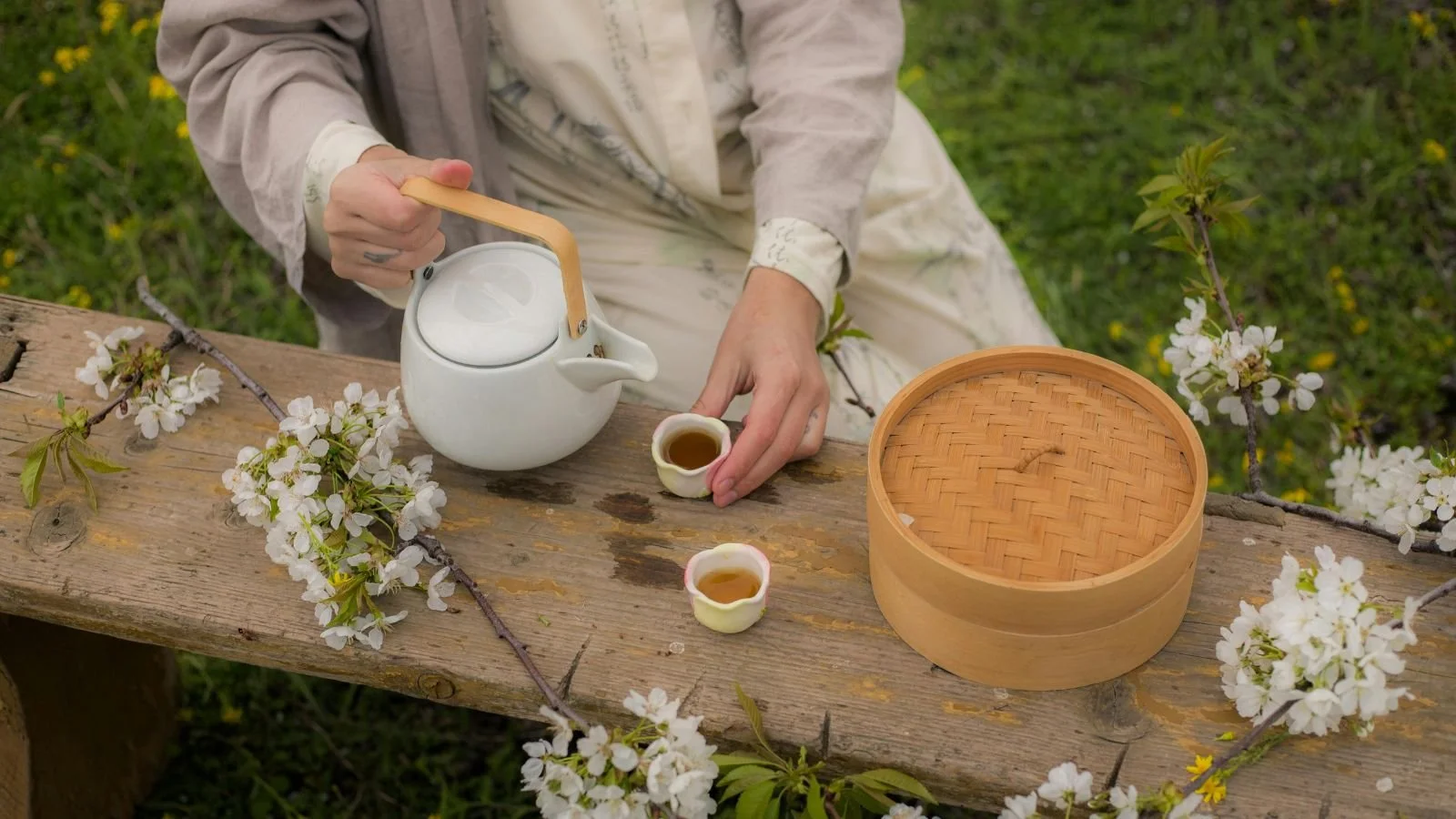 Person pouring tea from a white teapot into two small cups on a rustic wooden table surrounded by cherry blossoms in an outdoor setting.