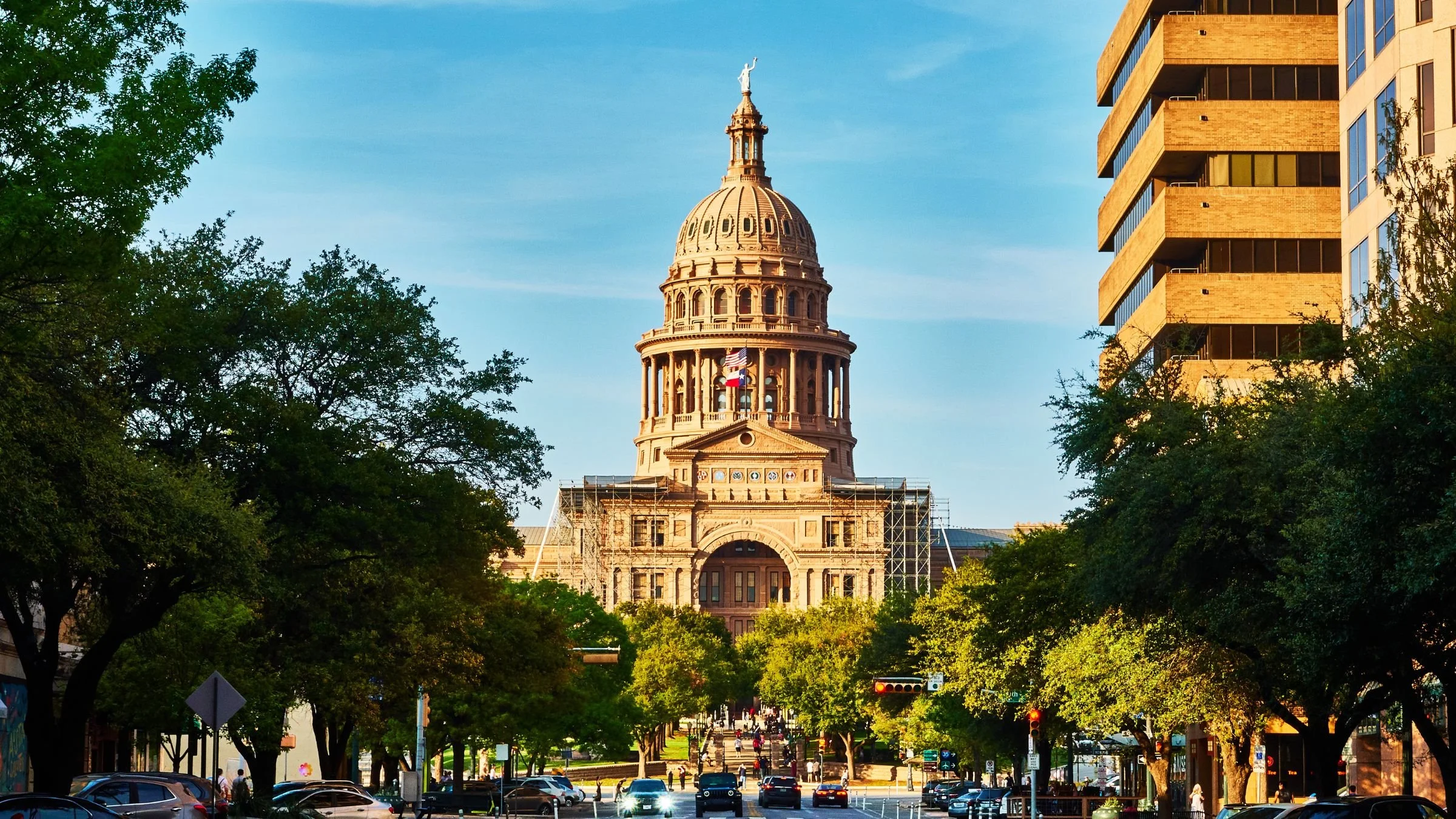 The Texas State Capitol building in Austin surrounded by trees and modern office buildings, with a parking lot and busy street in the foreground.