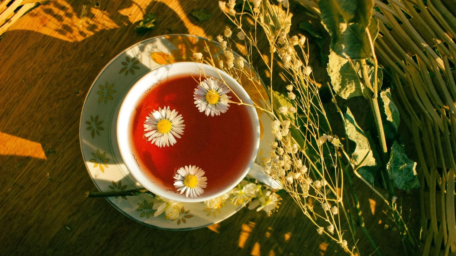 A cup of herbal tea with floating chamomile flowers, placed on a saucer with a floral design. The cup sits on a wooden surface surrounded by dried flowers and green leaves, with warm sunlight casting shadows.