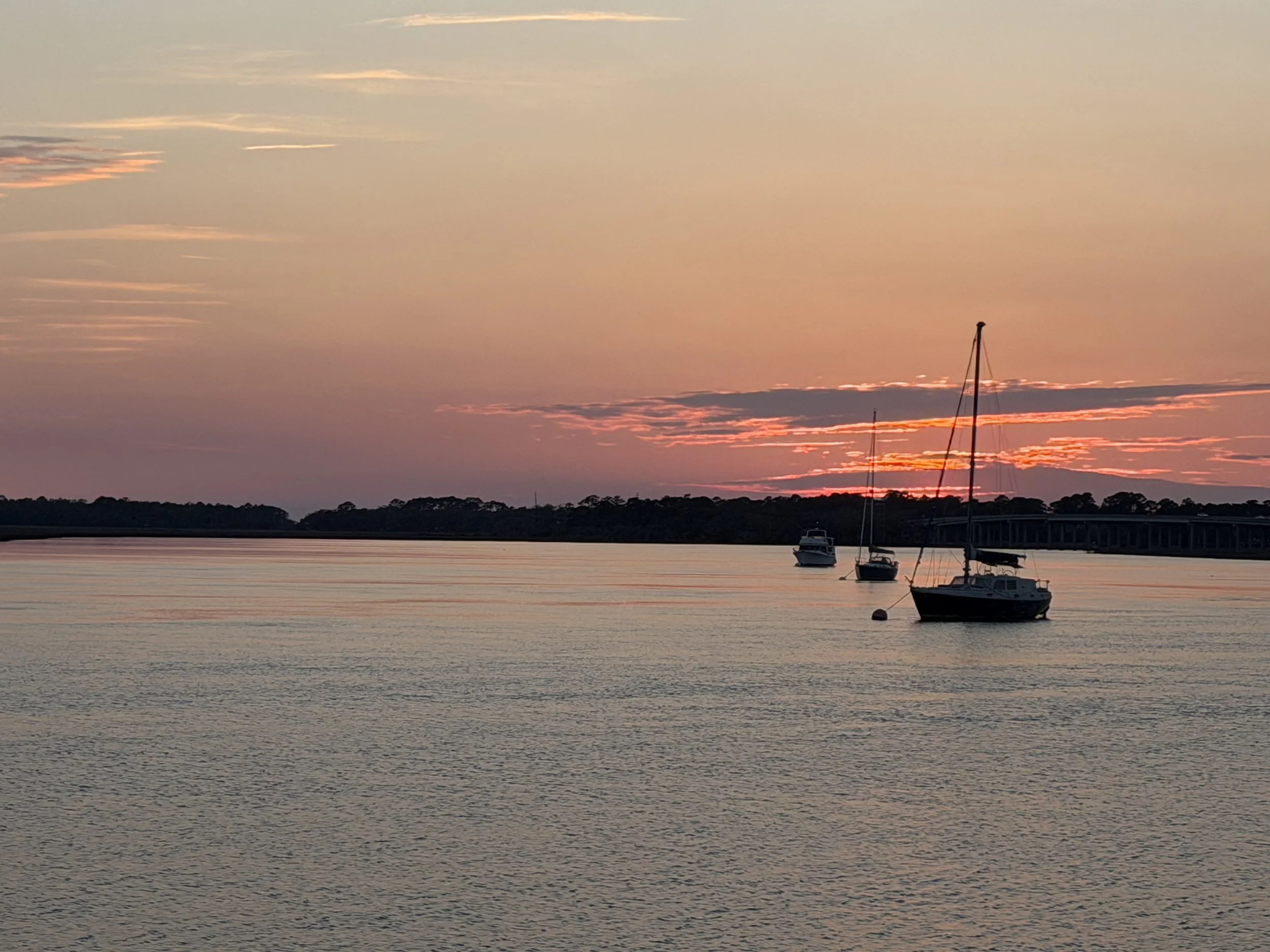 A sunset over a body of water with sailboats anchored near the shore, and a silhouetted landscape with trees and a bridge in the background.