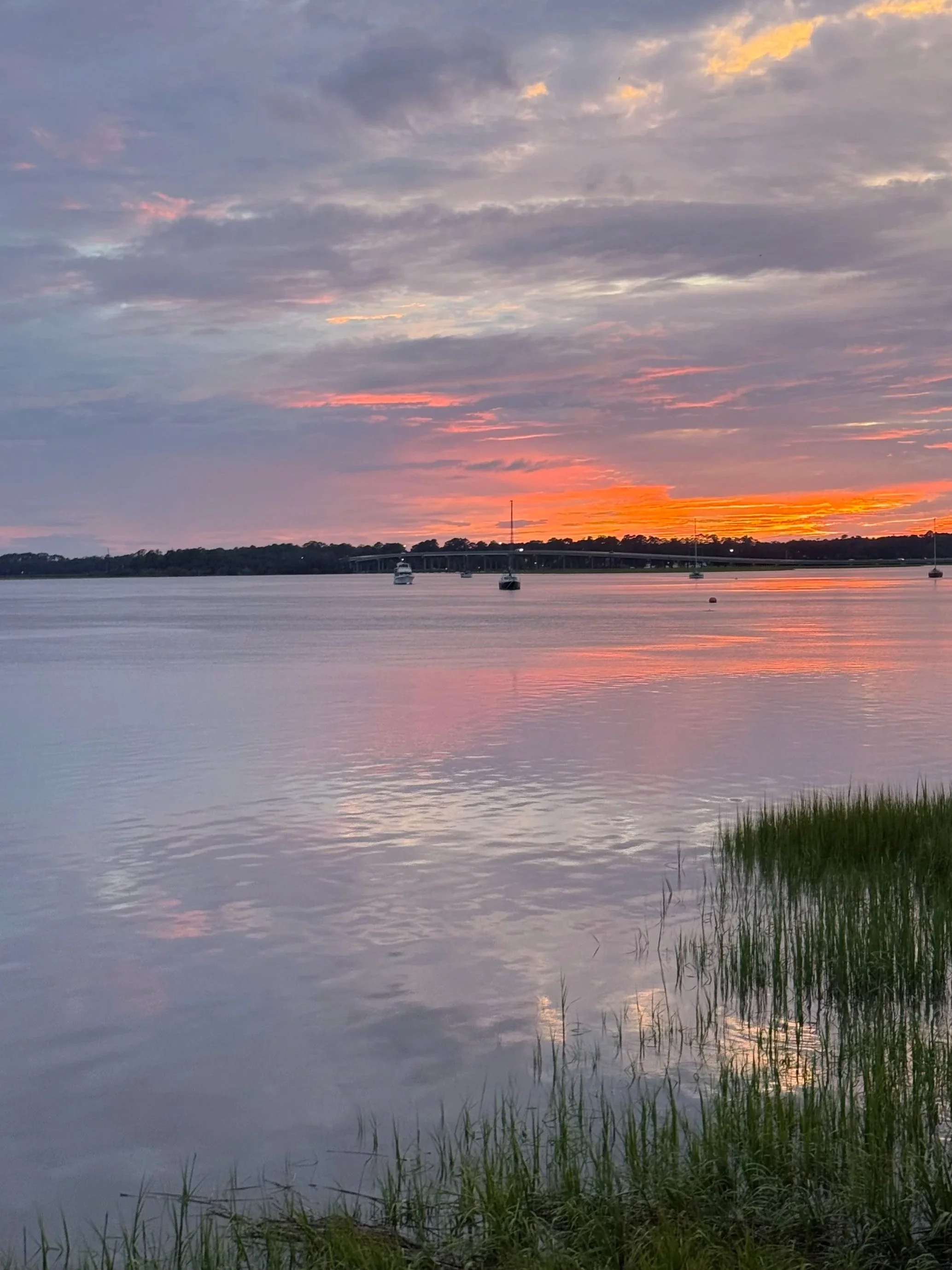 A serene sunset over a calm body of water with a few boats anchored, a distant bridge, and a partly cloudy sky with hues of orange, pink, and purple.