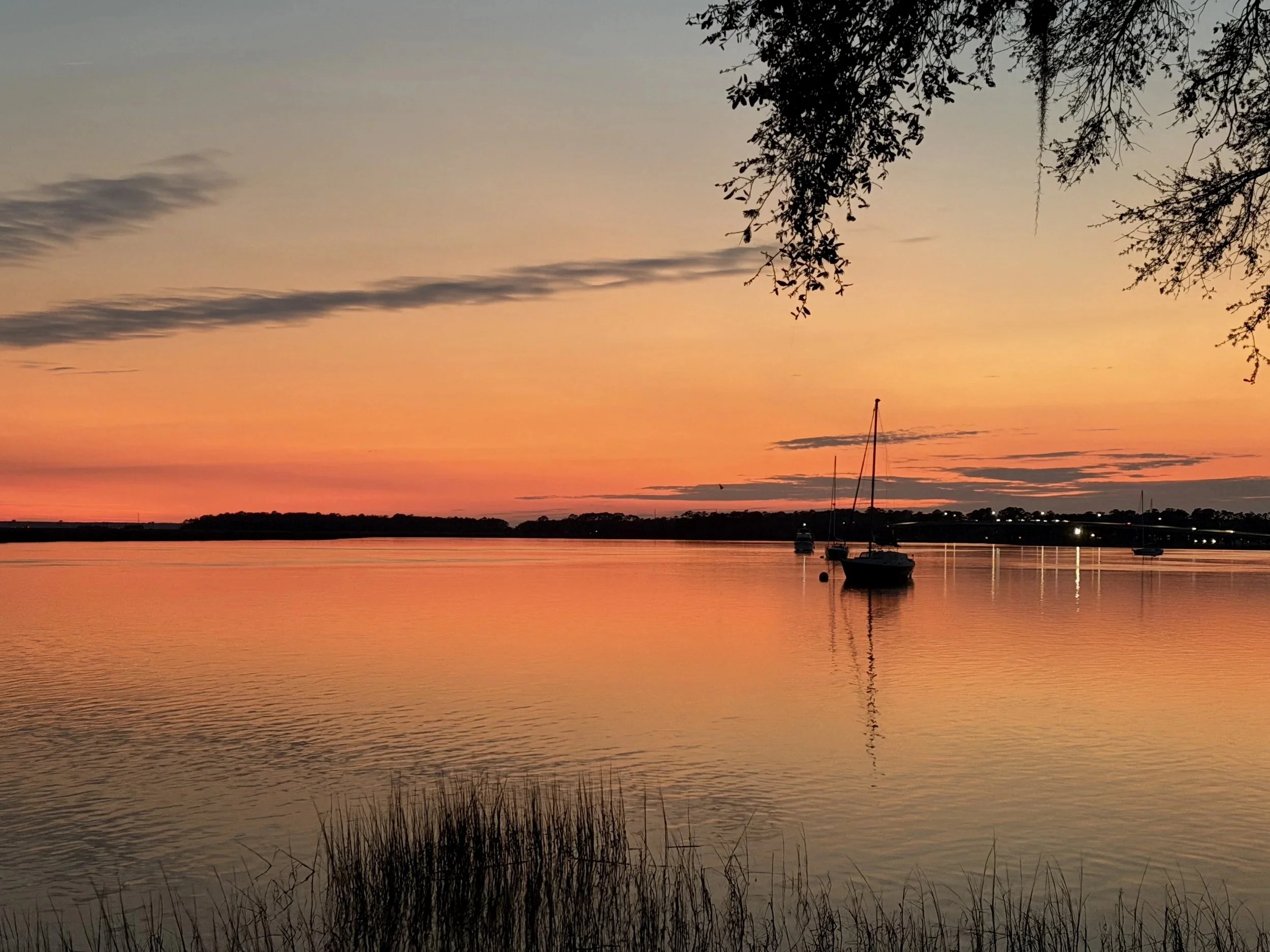 A peaceful sunset over a body of water with a orange and pink sky. Silhouettes of two sailboats are visible in the water, along with trees and land on the horizon.