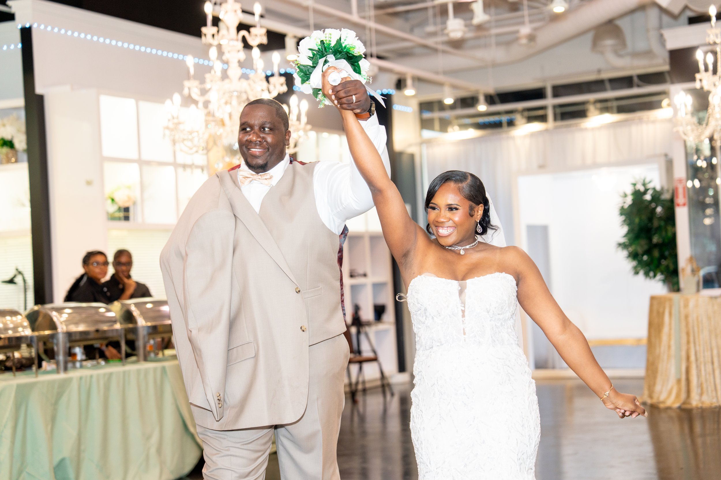 A newly married couple holding hands and raising a bouquet of flowers during their wedding reception, smiling in a decorated banquet hall with chandeliers.