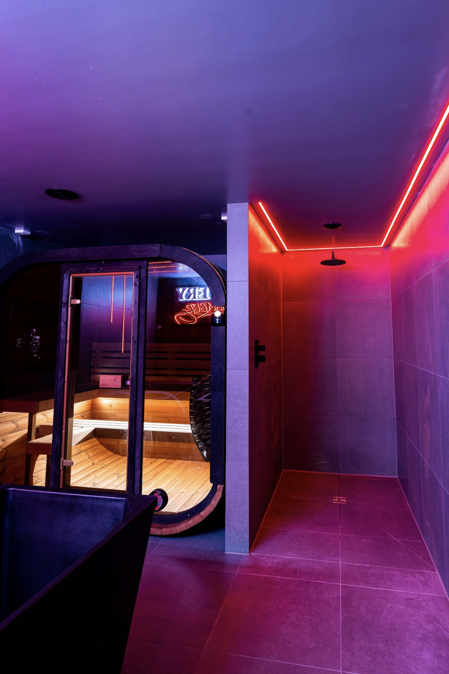 Indoor sauna room with glass door, wooden benches, and neon sign inside, adjacent to a shower area with red and purple LED lighting.