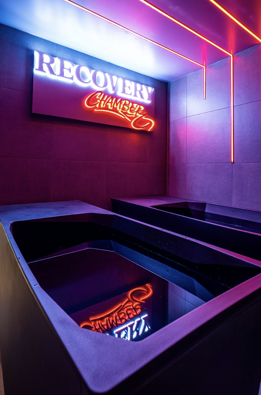 Neon sign reading "Recovery Chamber" on a purple wall with pink and purple neon lights, and two black hot tubs reflecting the sign.