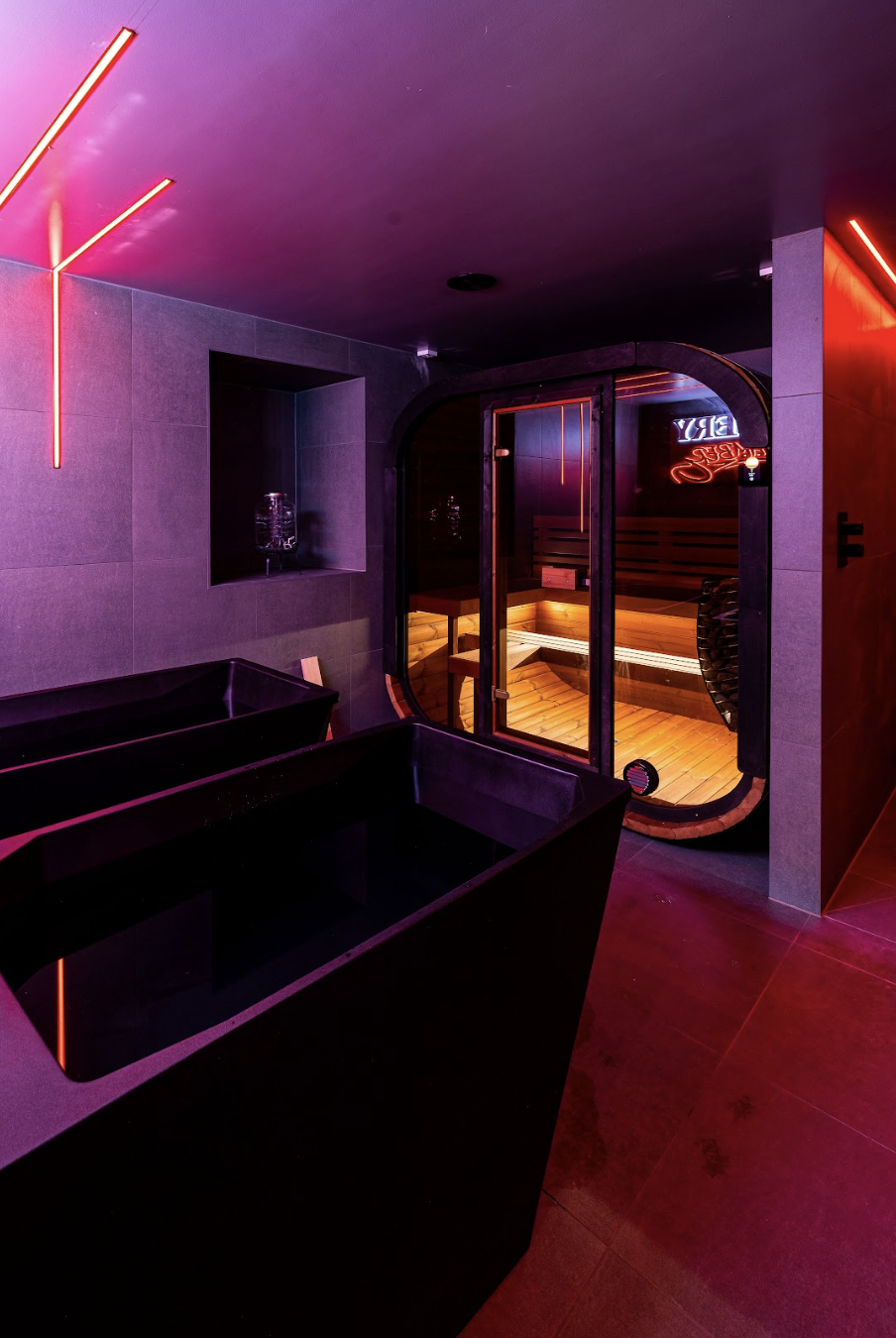 Interior of a modern spa room with LED lighting, a black tub, and a glass door leading to a wooden sauna with pink and purple lighting.