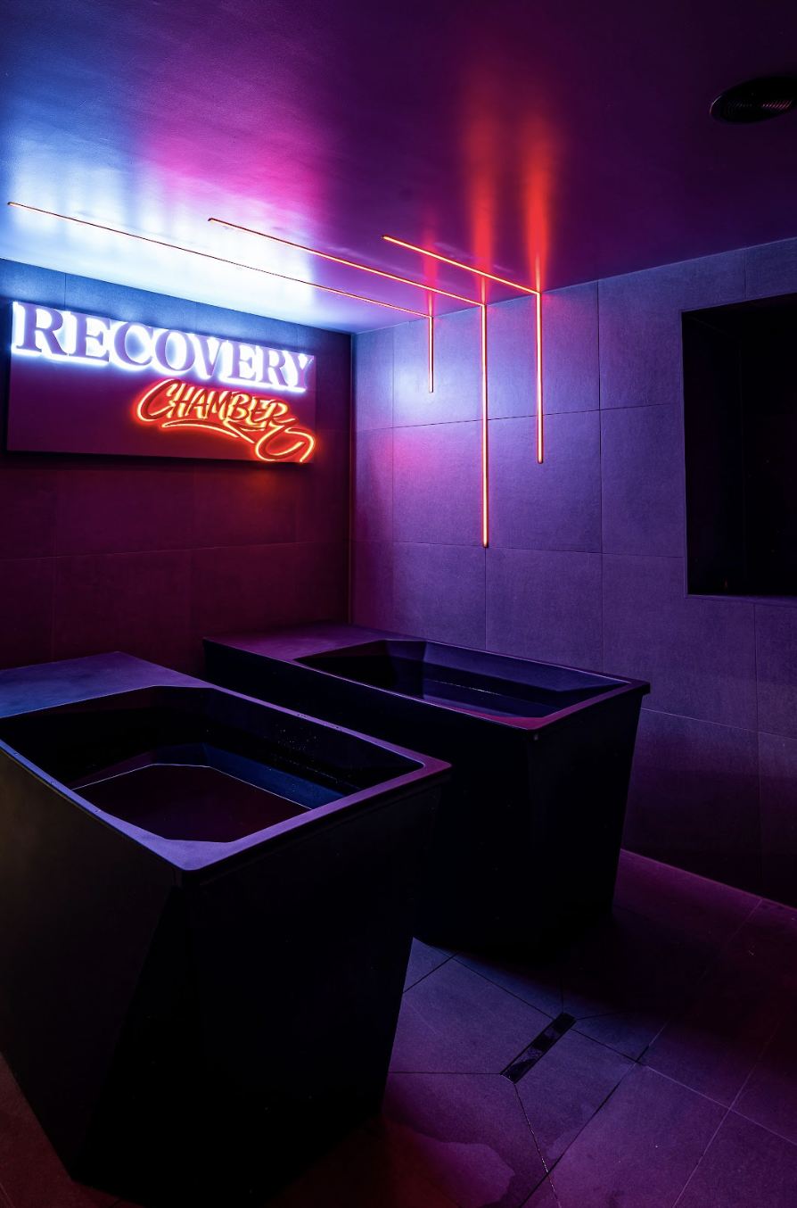 Dimly lit room with neon signs reading 'RECOVERY' and 'CHAMBER' on a wall, and two black bathtubs or hot tubs in front of the signs, illuminated by purple and red lighting.