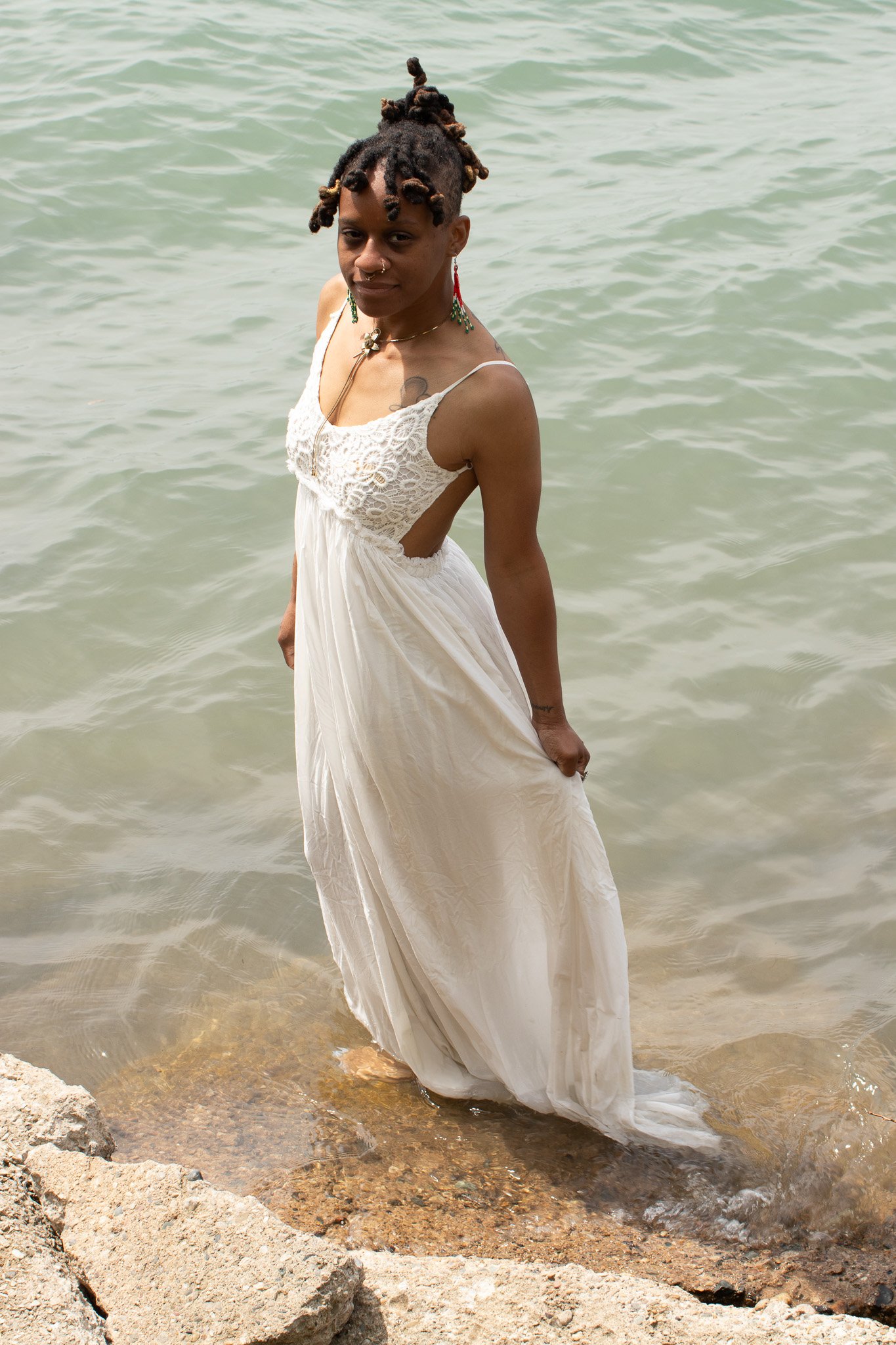 A woman with dark skin and dreadlocks standing in shallow water at a beach, wearing a white dress with lace details, earrings, and a nose ring.