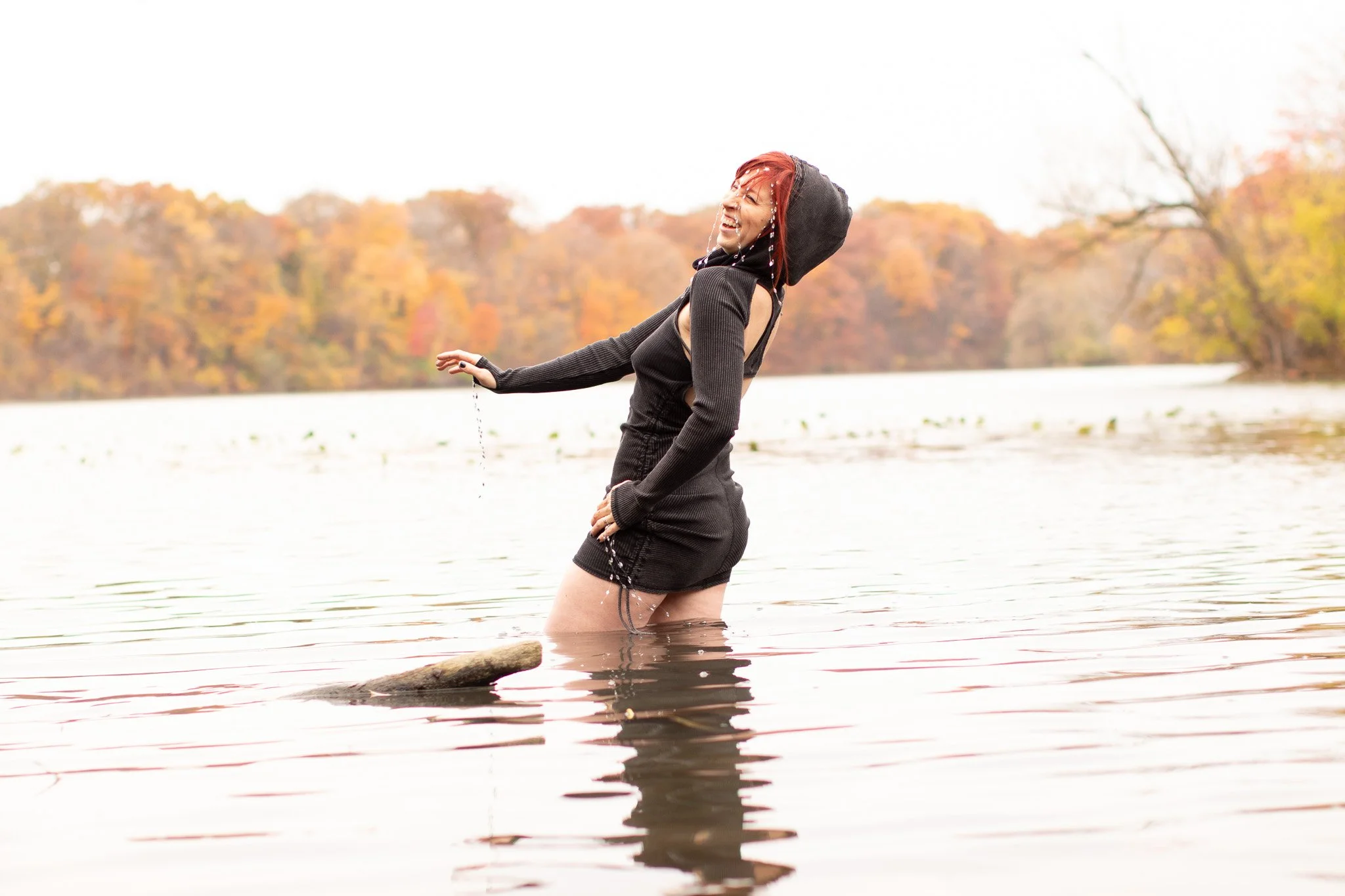 A woman with red hair and a black hoodie, smiling and splashing water while standing in a lake with autumn trees in the background.