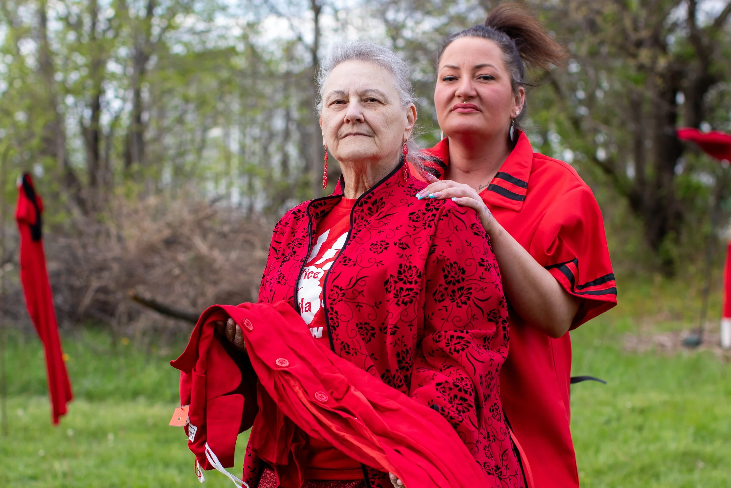 Two women outdoors, one older with gray hair in a red and black patterned jacket, and one younger with brown hair in a high ponytail, in red clothing, standing in a wooded area.
