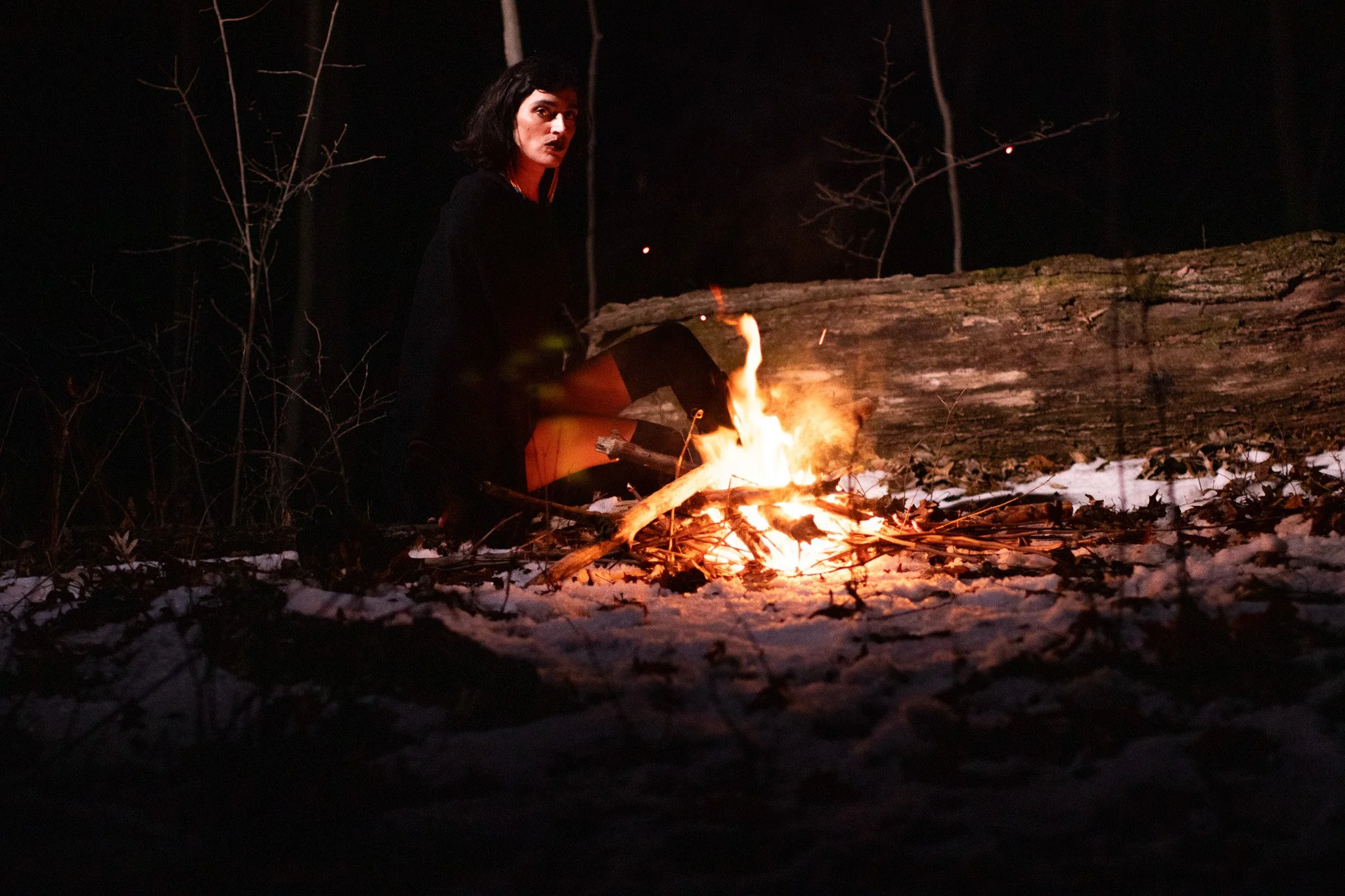 A woman sitting by a campfire at night in a wooded area. She has short dark hair and is wearing a black dress. The fire is burning on the snowy ground, illuminating her face and surroundings.