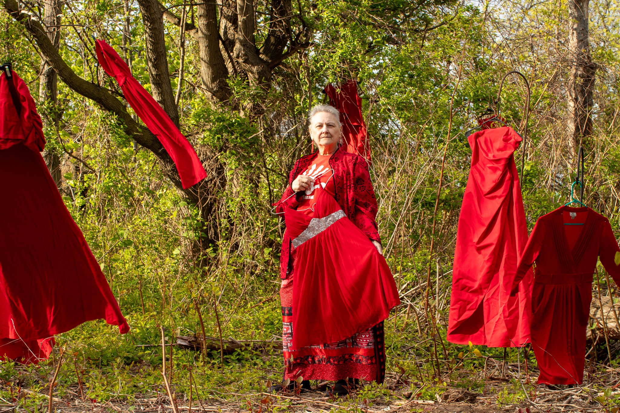 An elderly woman standing in a wooded area holding a red dress. Red dresses are hanging on hangers and are draped over tree branches around her.
