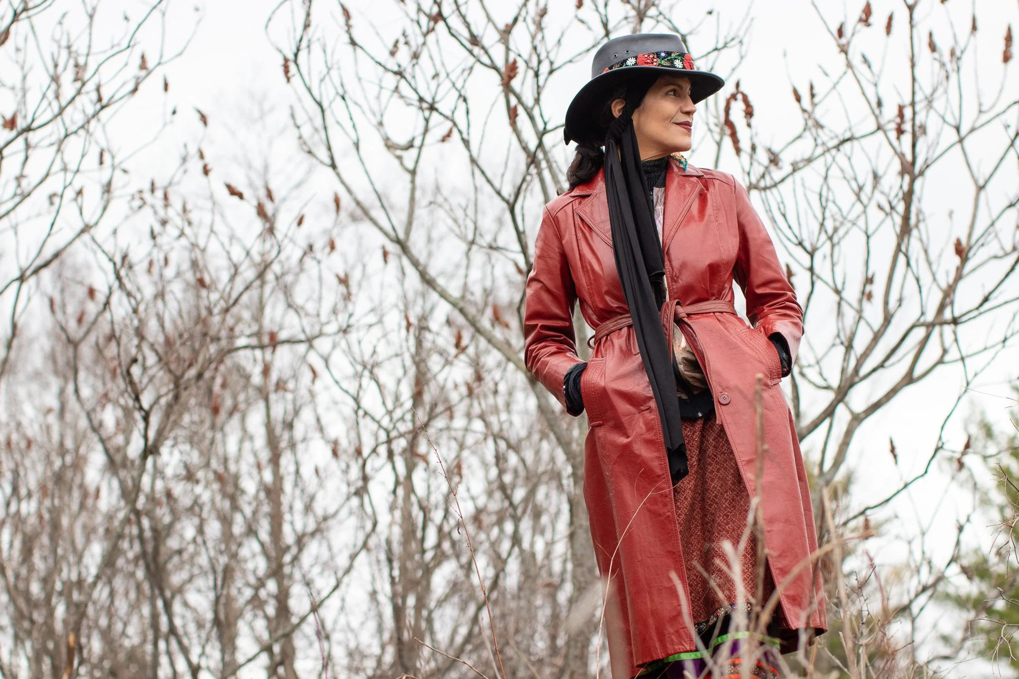 A woman wearing a red leather jacket and a black hat with floral embroidery stands outdoors among sumac trees on a cloudy day.