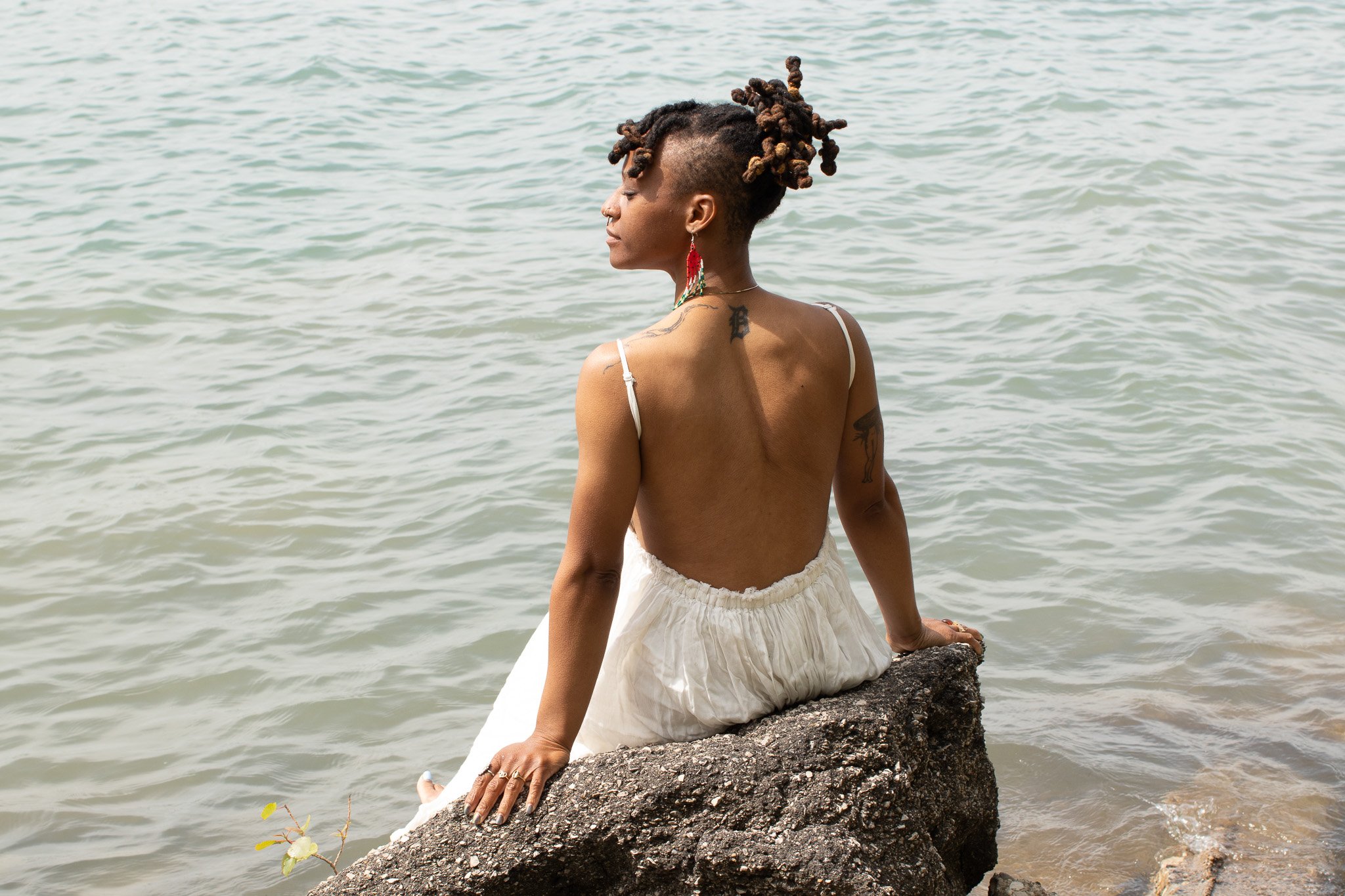A woman with short dreadlocks and tattoos wearing earrings and a white dress, sitting on a rock at the water's edge with eyes closed.