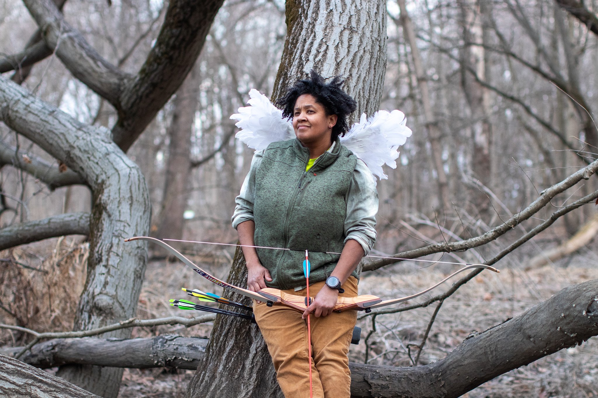 A woman standing on a fallen tree in a forest with leafless trees, wearing a green jacket, tan pants, and angel wings, holding a bow and arrows.