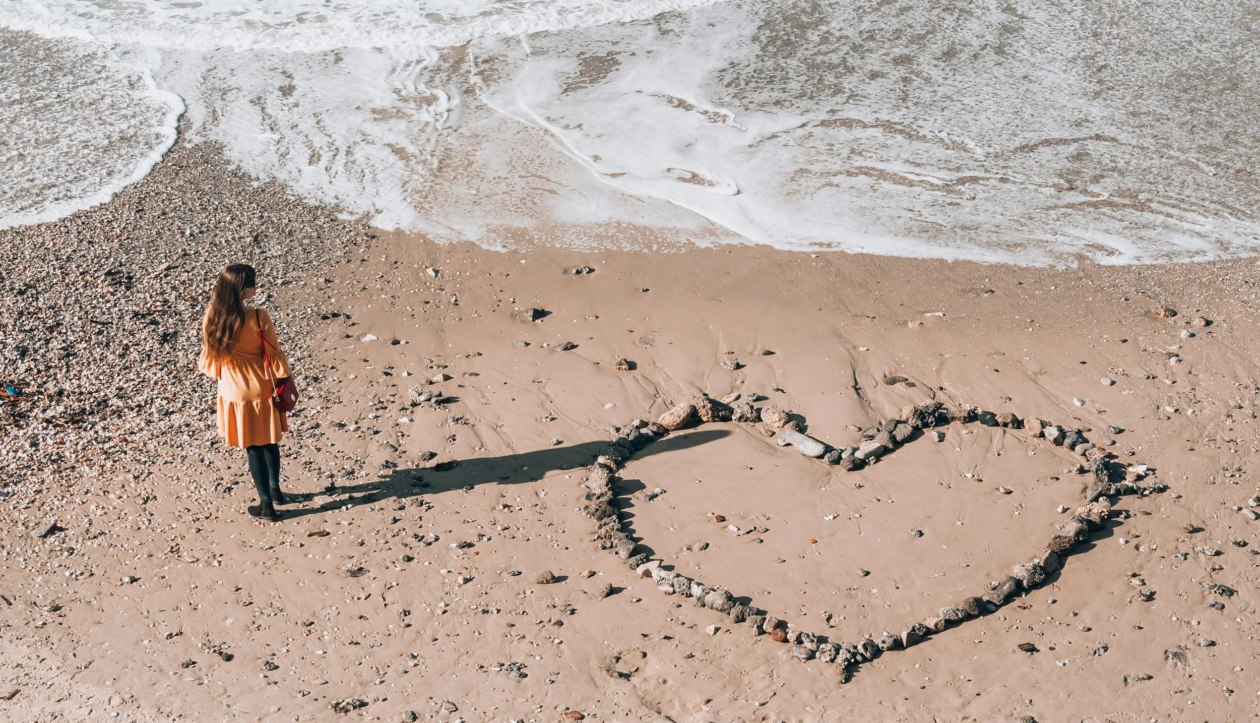 A woman with long hair wearing an orange dress and black leggings stands on a sandy beach, facing the ocean, next to a heart-shaped outline made of rocks.