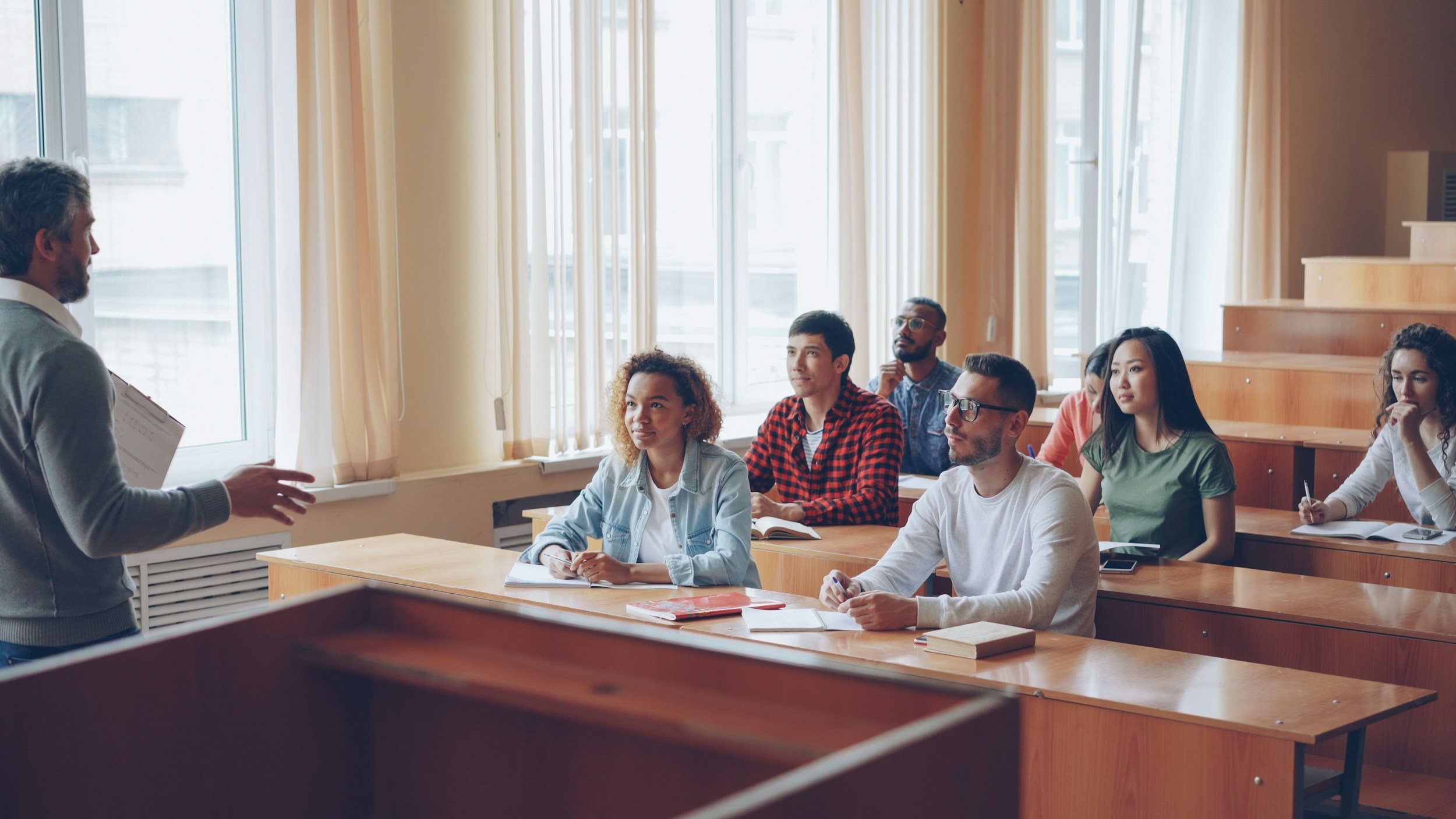 A classroom with students sitting at desks listening to a teacher who is standing and speaking at the front of the room.