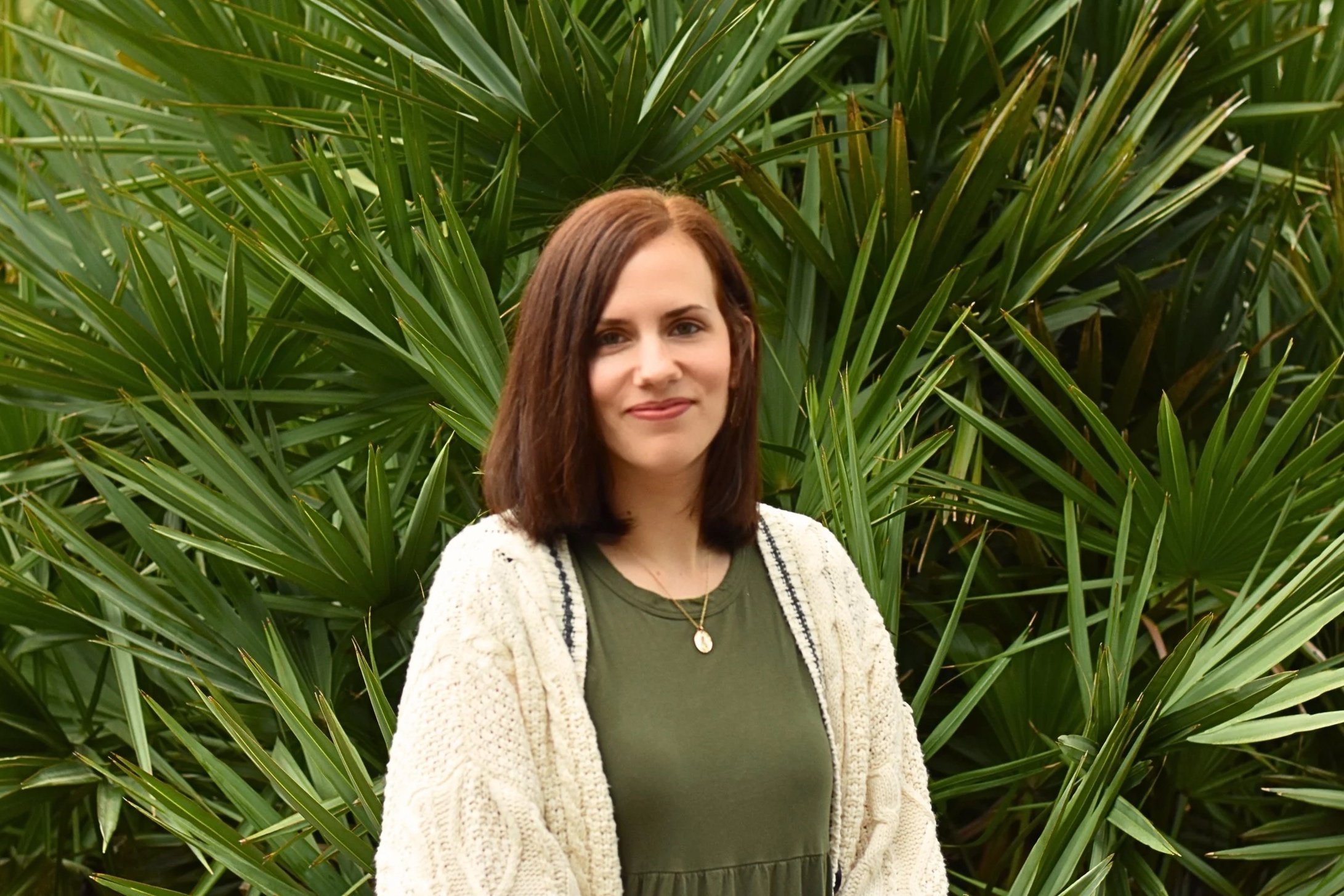 A woman with brown hair standing in front of large green leafy plants, wearing a green top, a beige cardigan, and a necklace.
