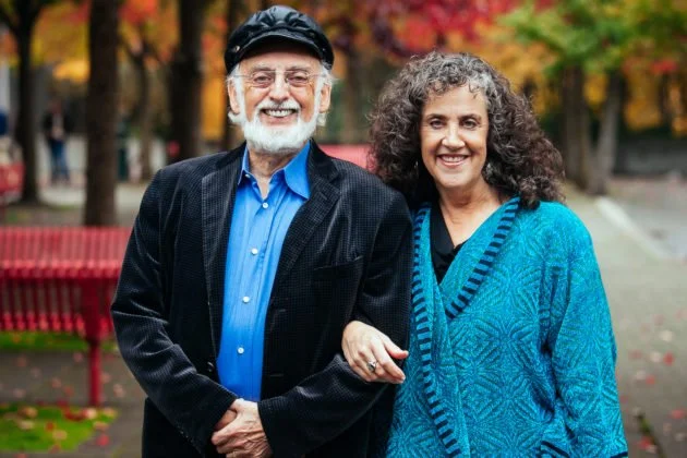 An elderly man and woman smiling and walking together in a park with autumn trees in the background.