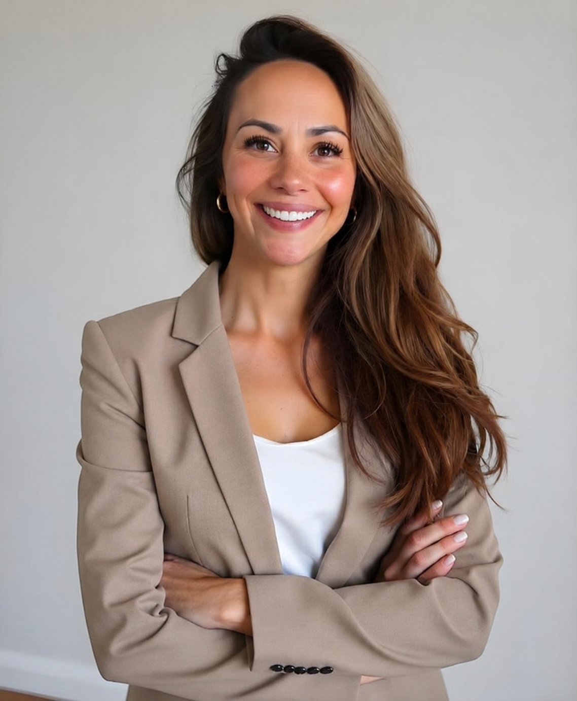 A smiling woman with long, wavy brown hair, wearing a beige blazer over a white top, standing with arms crossed against a plain light gray background.