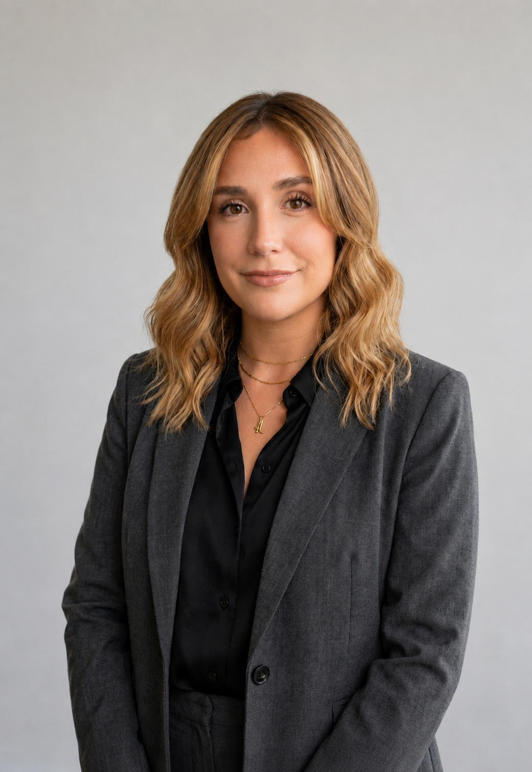 A woman with shoulder-length wavy blonde hair, wearing a dark gray blazer over a black shirt, standing against a plain light gray background.