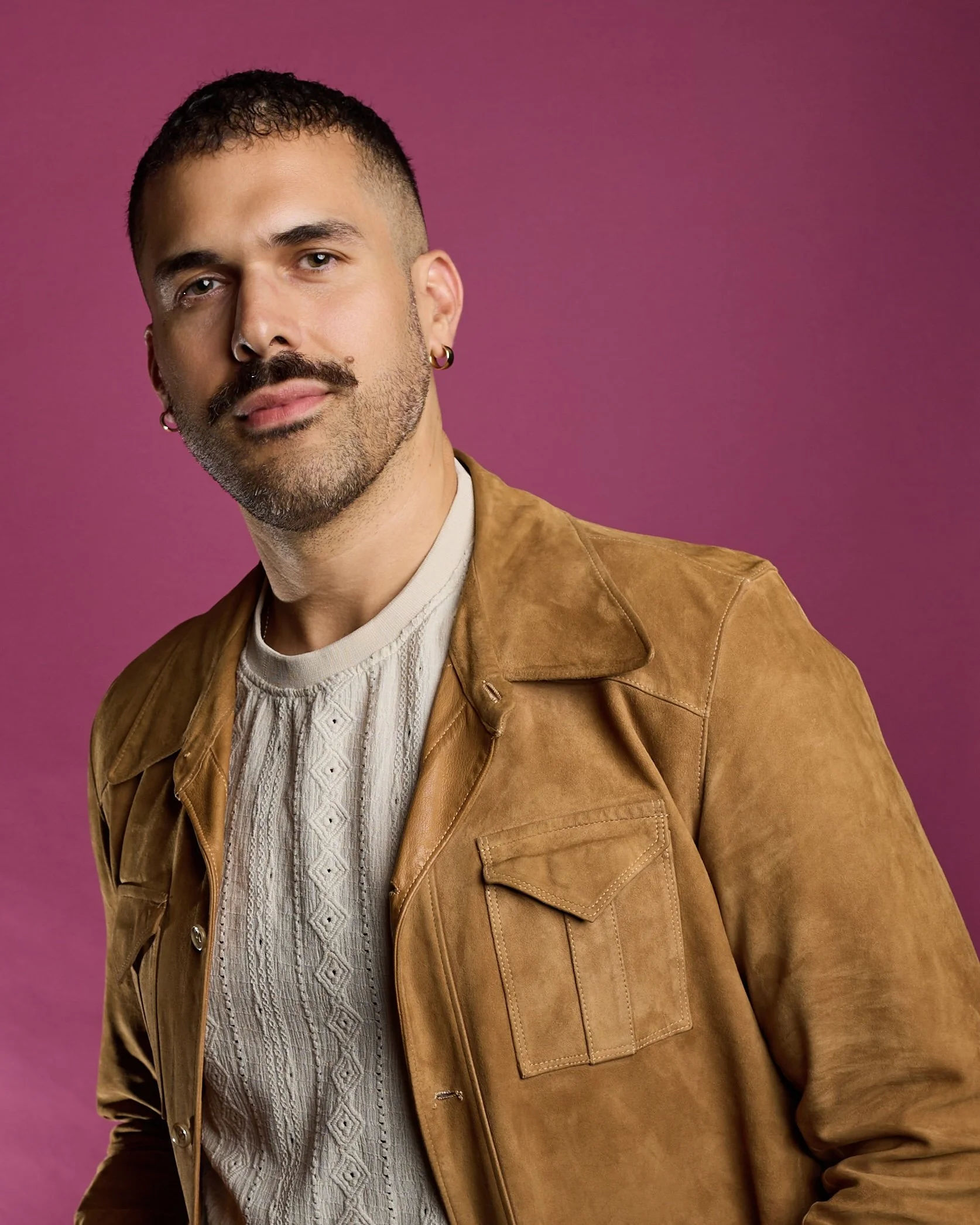 A young man with short dark hair, a mustache, and goatee, wearing a brown leather jacket over a cream-colored cable knit sweater, posing against a pink background.