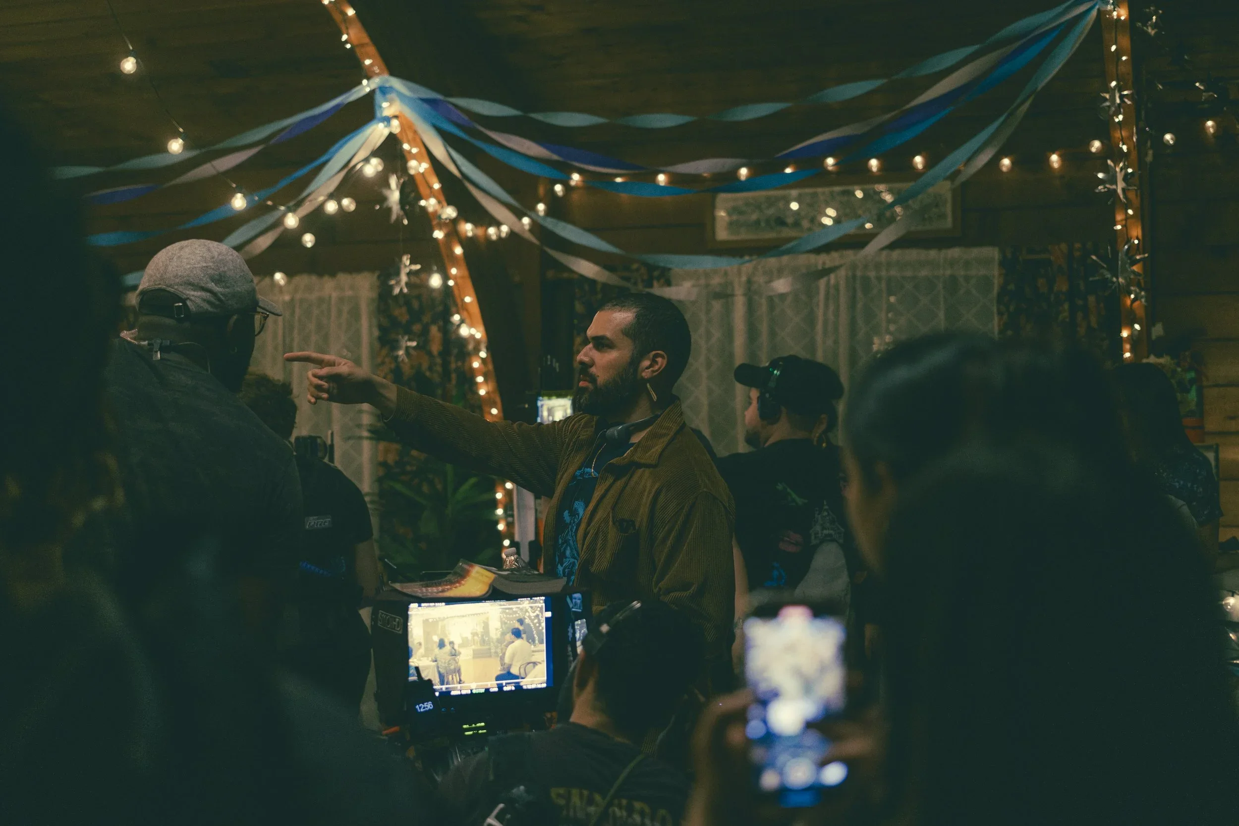 A man with a beard points towards a display monitor at a gathering or party, with people around and string lights and blue streamers hanging from the ceiling.