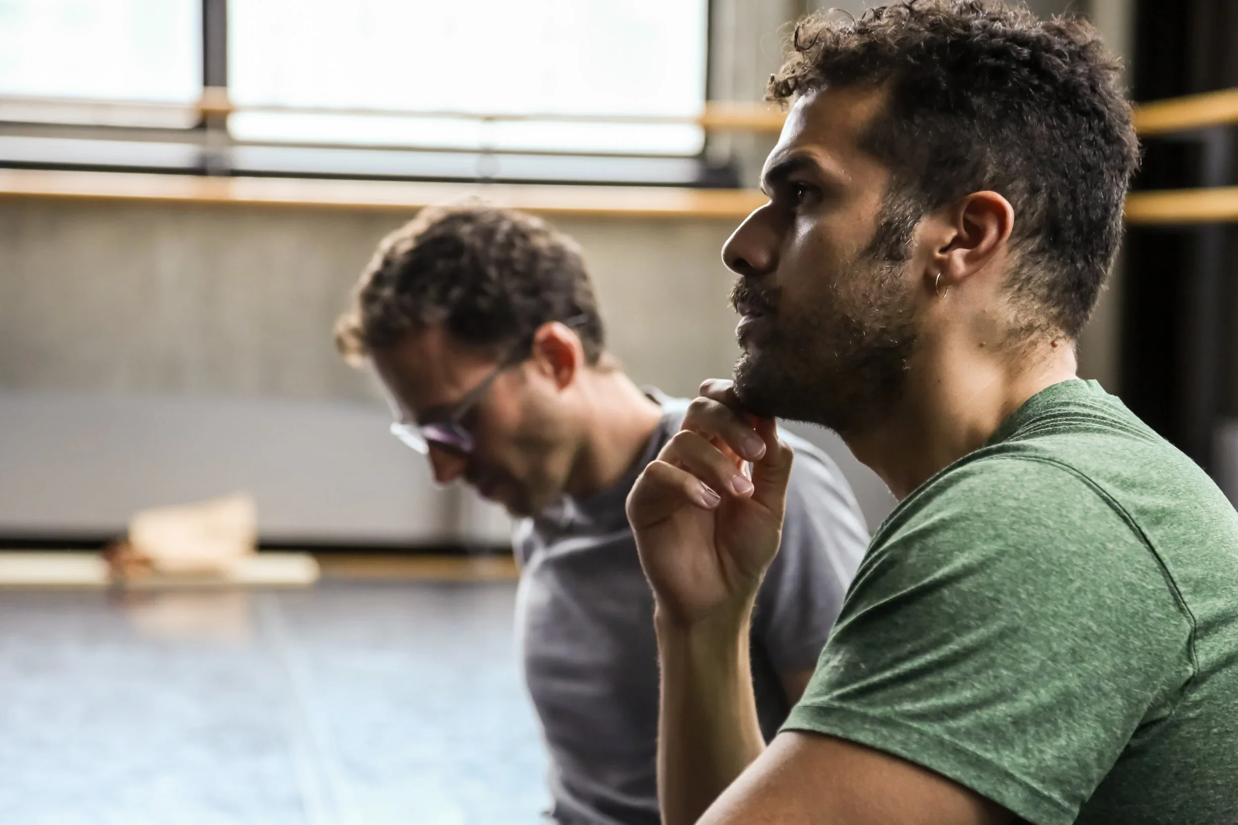 Two men sitting in profile, one with a thinking pose and hand on chin, in an indoor setting with large windows in the background.