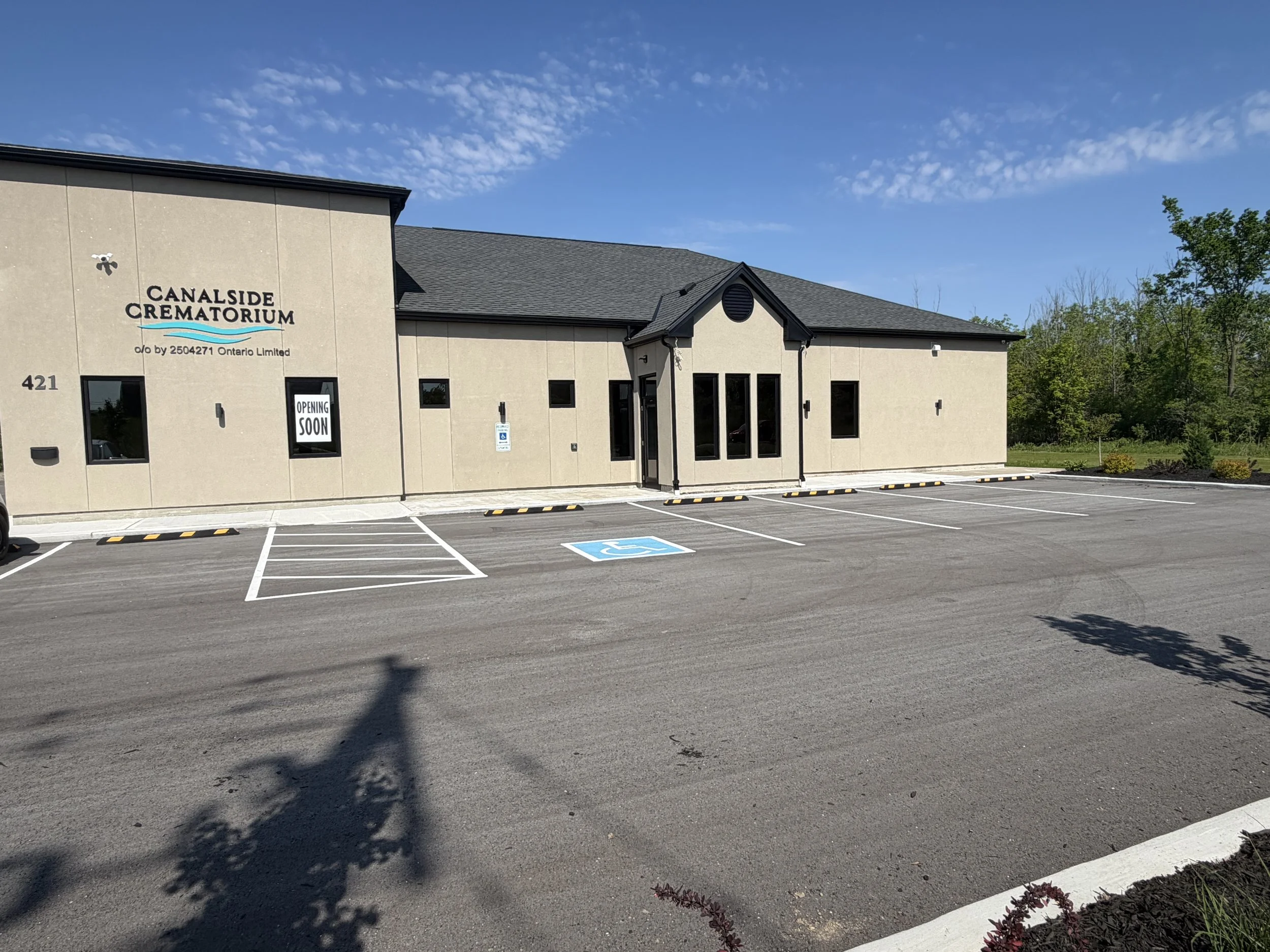 Exterior view of Canalside Crematorium building with parking lot, handicap parking space, and an 'opening soon' sign, under a blue sky with scattered clouds.