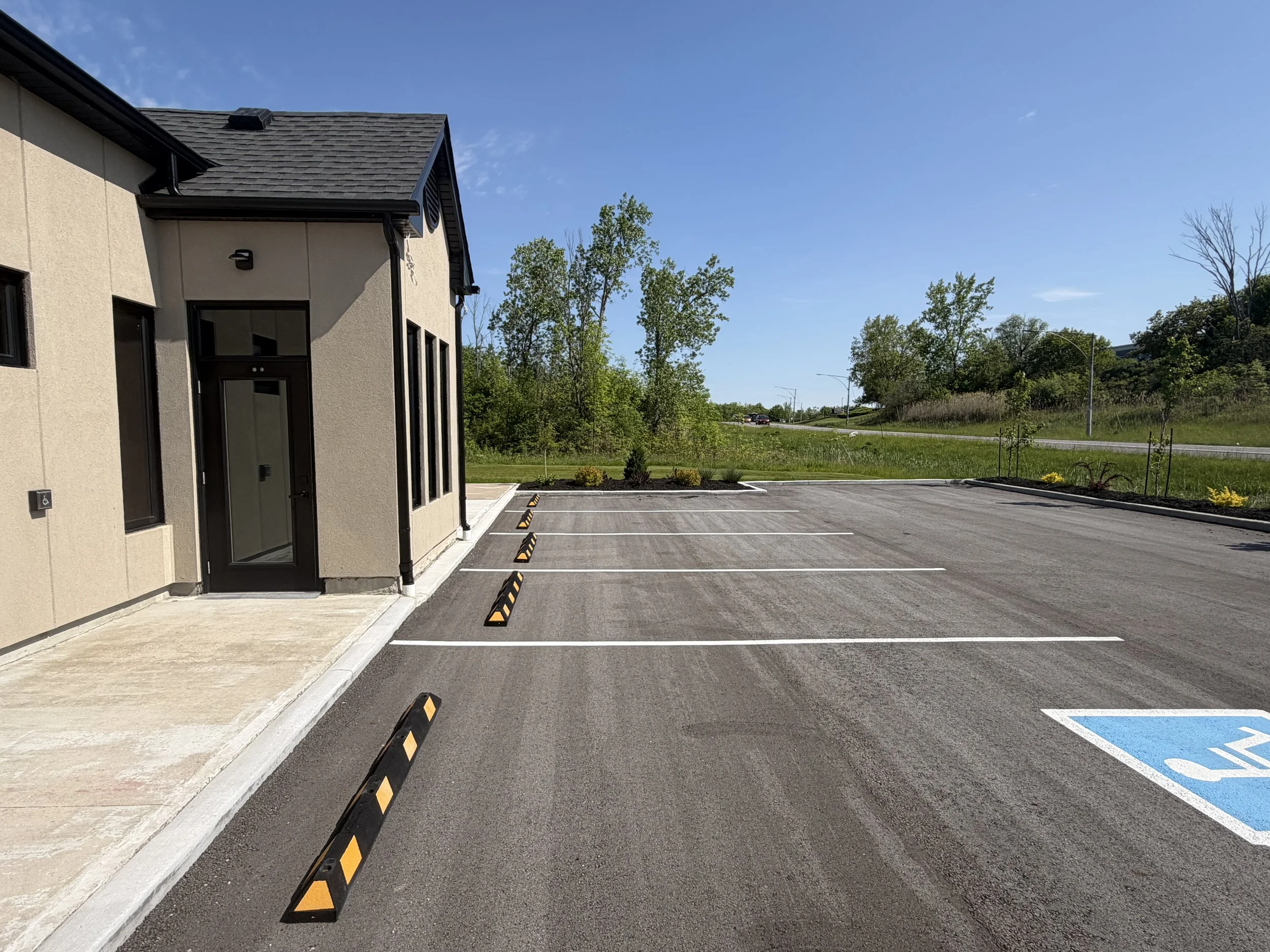 Empty parking lot with designated handicapped parking spot, a building on the left, and a grassy area with trees and clear blue sky in the background.