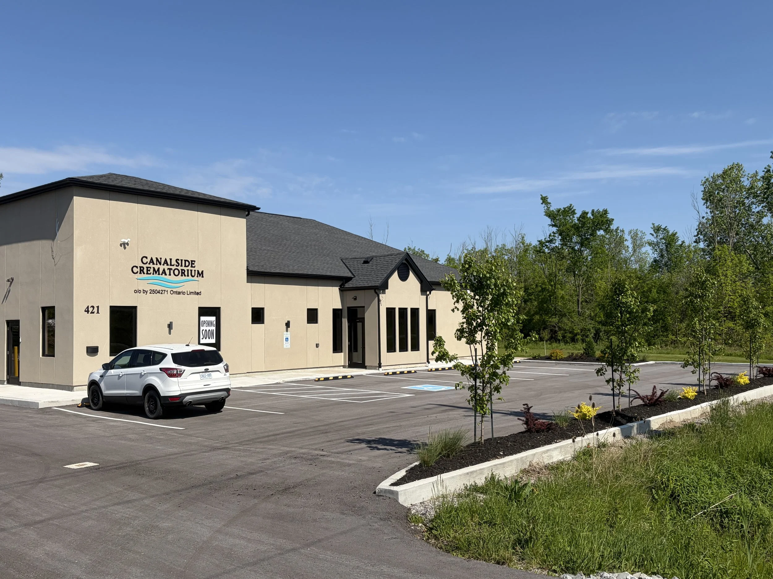 Exterior of a building with a sign that reads 'Canalside Crematorium' and a parking lot with a white SUV, surrounded by small landscaped trees and bushes, under a blue sky with some clouds.