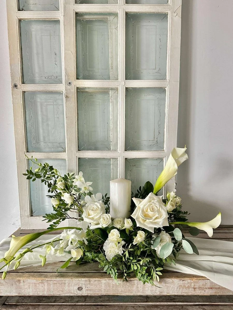 A floral arrangement of white roses, calla lilies, and greenery with a white candle in the center, placed on a wooden surface in front of a glass-paned white window.