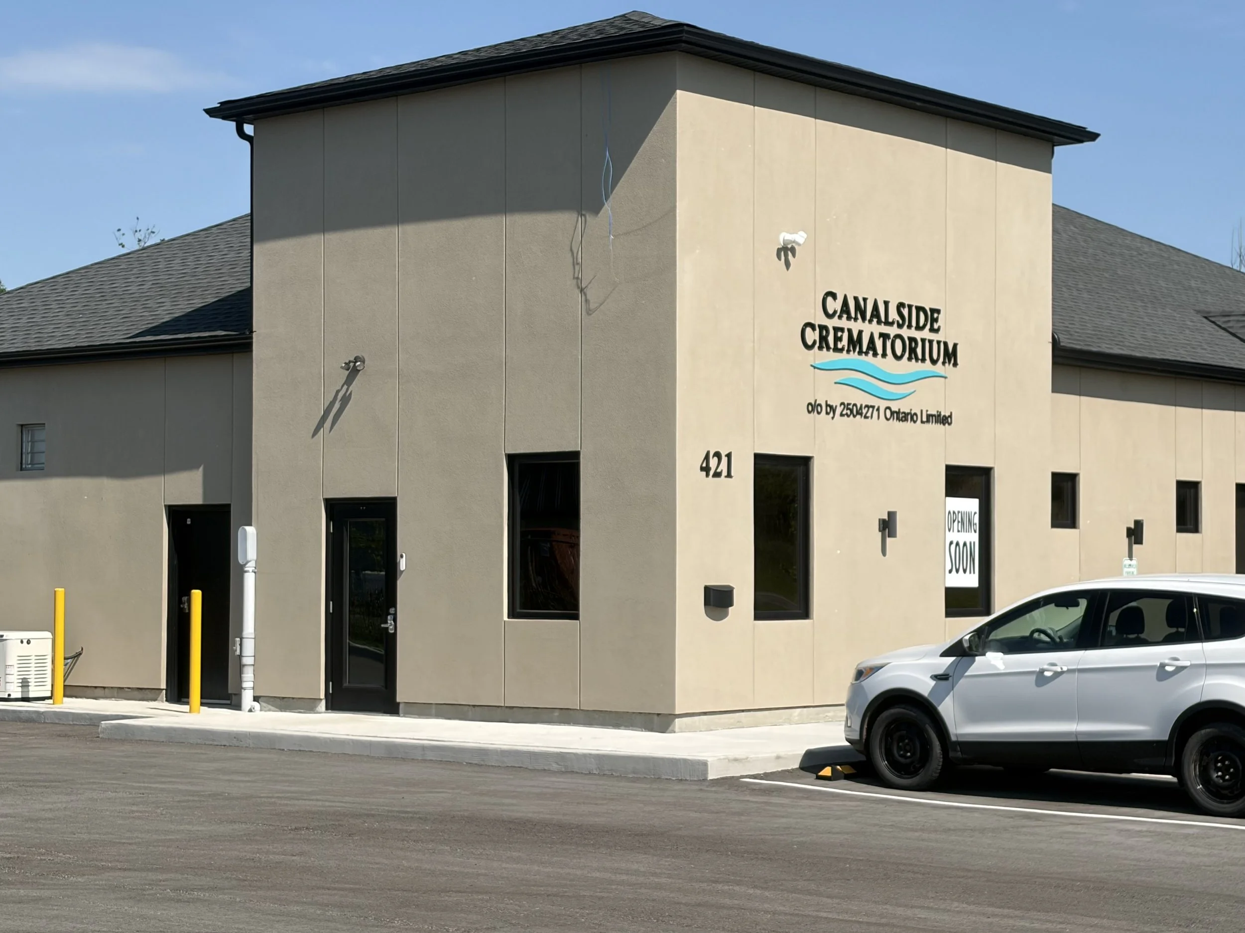 The image shows a beige building with the sign 'Canalside Crematorium' on it, located at 421. There are several parking spots in front, with a white car parked and yellow bollards near the entrance. Part of the building's roof and a blue sky are visible.