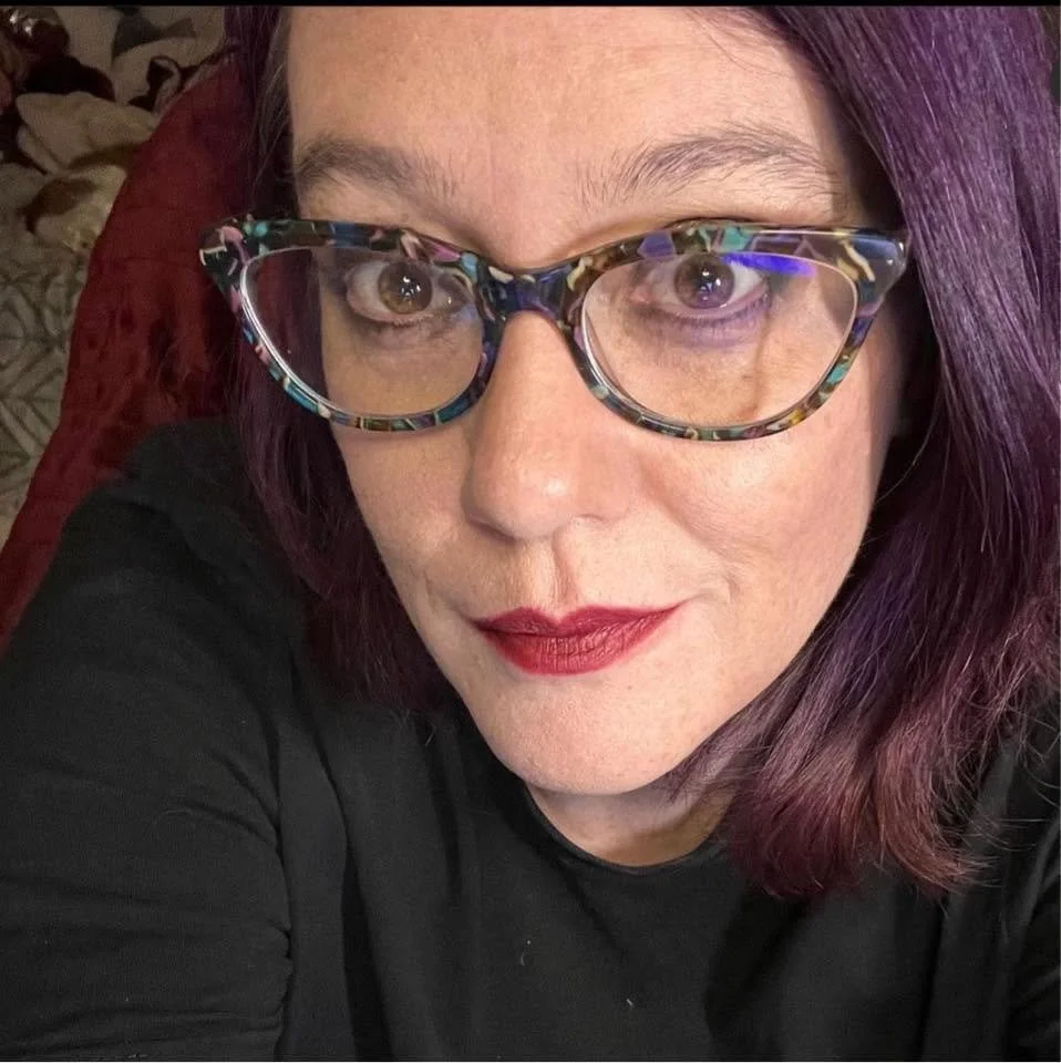 Close-up of a woman with purple hair, wearing colorful glasses and red lipstick, looking at the camera.