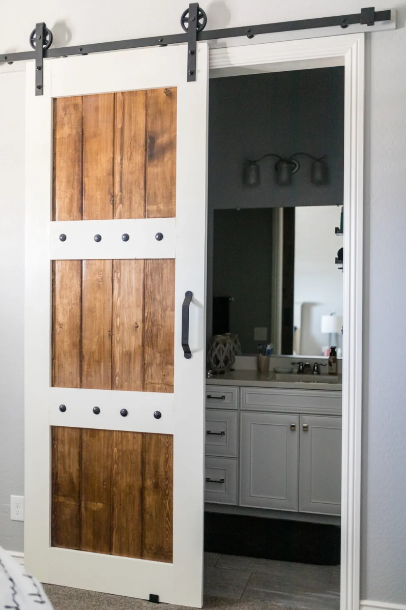 A white sliding barn door with three panels of natural wood and black hardware, partially open, revealing a bathroom with a large mirror, vanity, and dark wall.