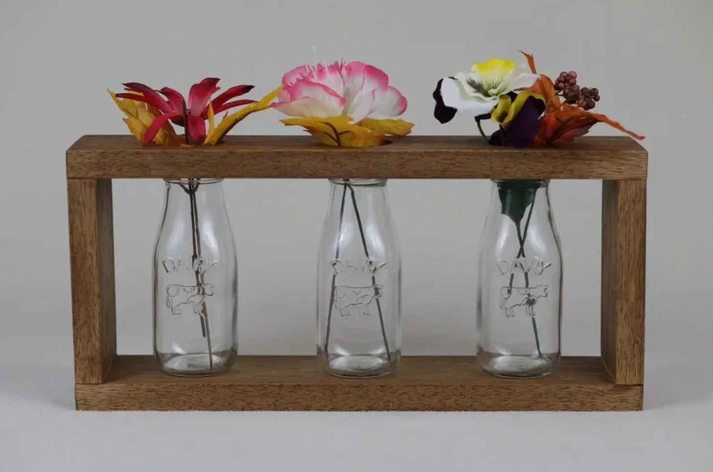 Three small glass bottles with flowers, mounted on a wooden frame, with flowers in various colors including red, pink, yellow, white, and purple.