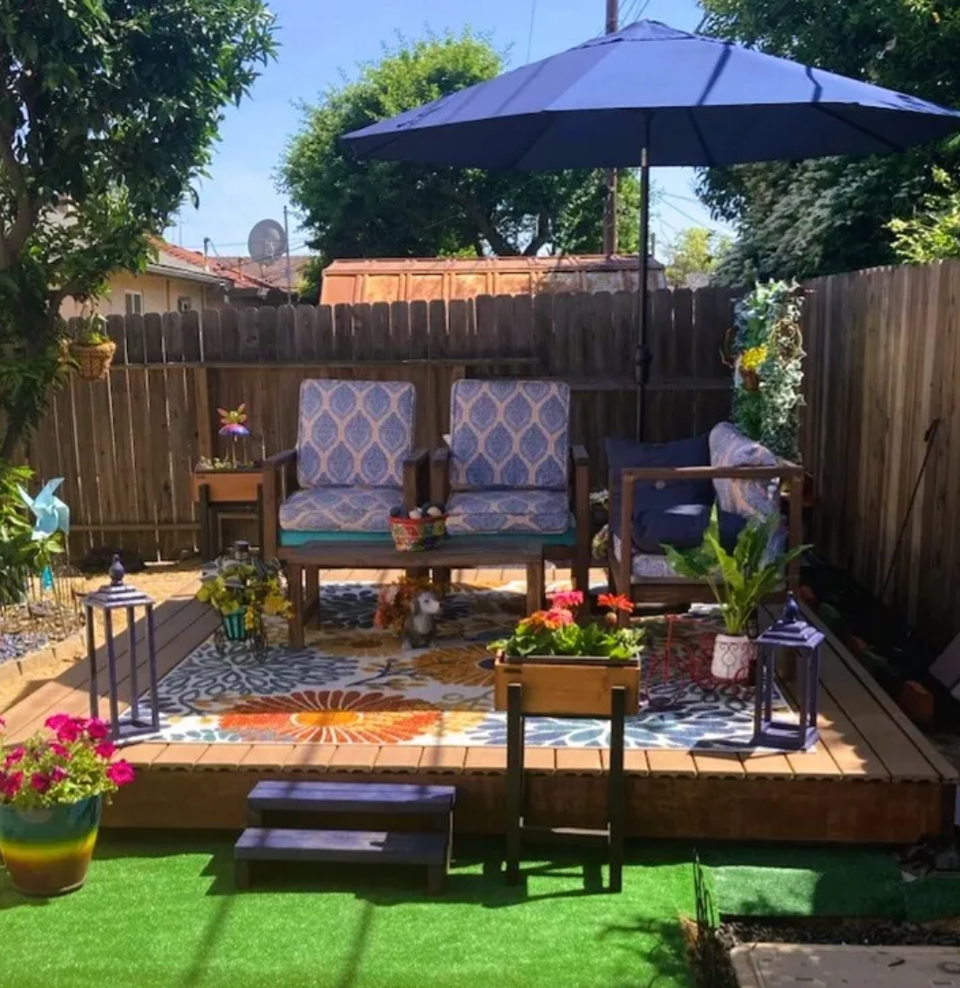 A cozy backyard patio with a wooden deck, patterned outdoor furniture, a large blue patio umbrella, colorful potted plants, lanterns, and decorative garden elements.