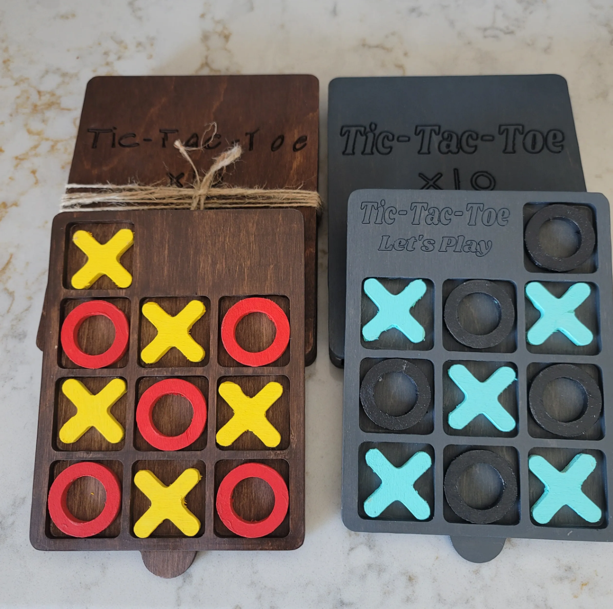 Colorful tic-tac-toe game boards on a marble surface. The left board has yellow X's and red O's, and the right black board has light blue X's and black O's. Both boards are labeled 'Tic-Tac-Toe', with the right board also saying 'Let's Play'.