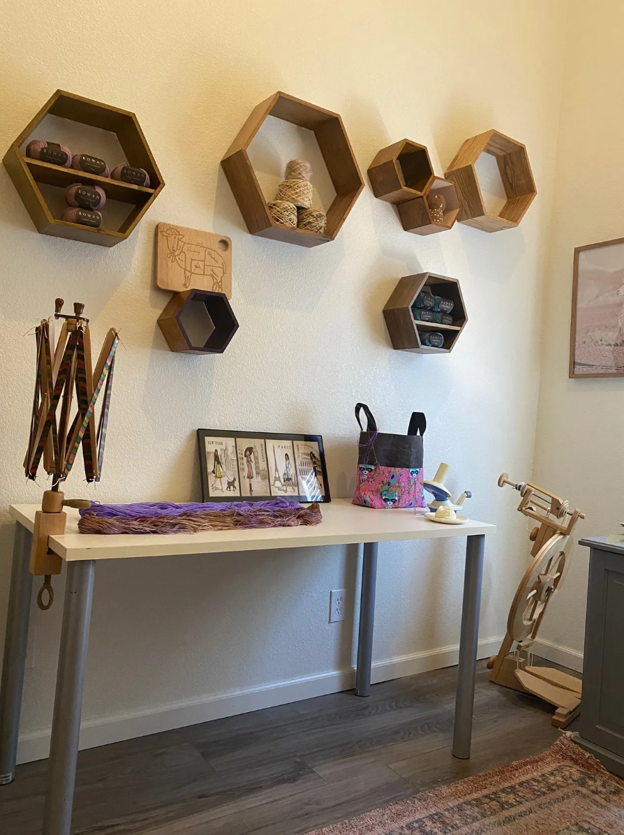A craft or sewing room with a white table holding a colorful bag, framed pictures, and craft supplies. Wooden hexagon shelves on the wall display yarn, thread, and small containers. A wooden spinning reel is in the corner next to a light-colored wall