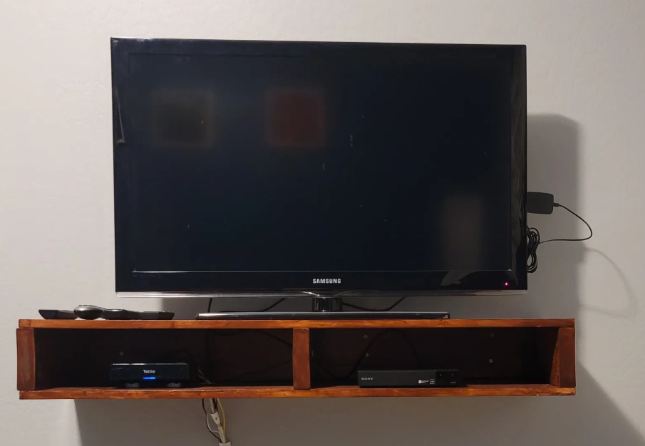 A flat-screen Samsung television mounted on a wall, sitting above a small wooden shelf with electronic devices and remote controls.