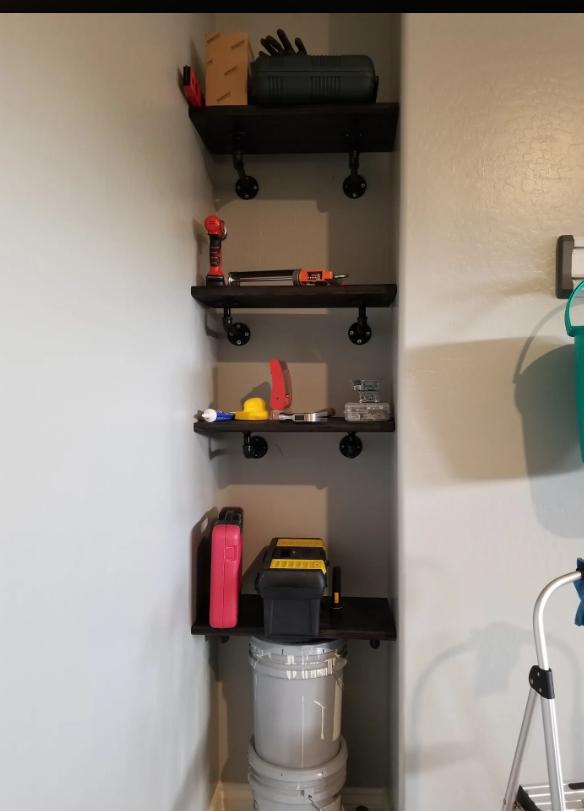 Narrow wall-mounted shelving unit with five shelves holding various tools and supplies, including power drills, a tape measure, a flashlight, and storage containers, with a bucket below the bottom shelf.