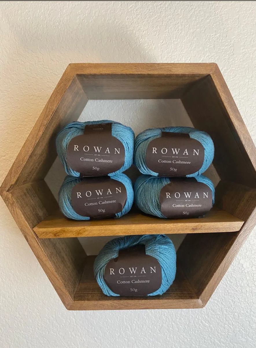 Hexagonal wooden shelf mounted on wall, holding five skeins of Rowan cotton cashmere yarn in shades of blue.