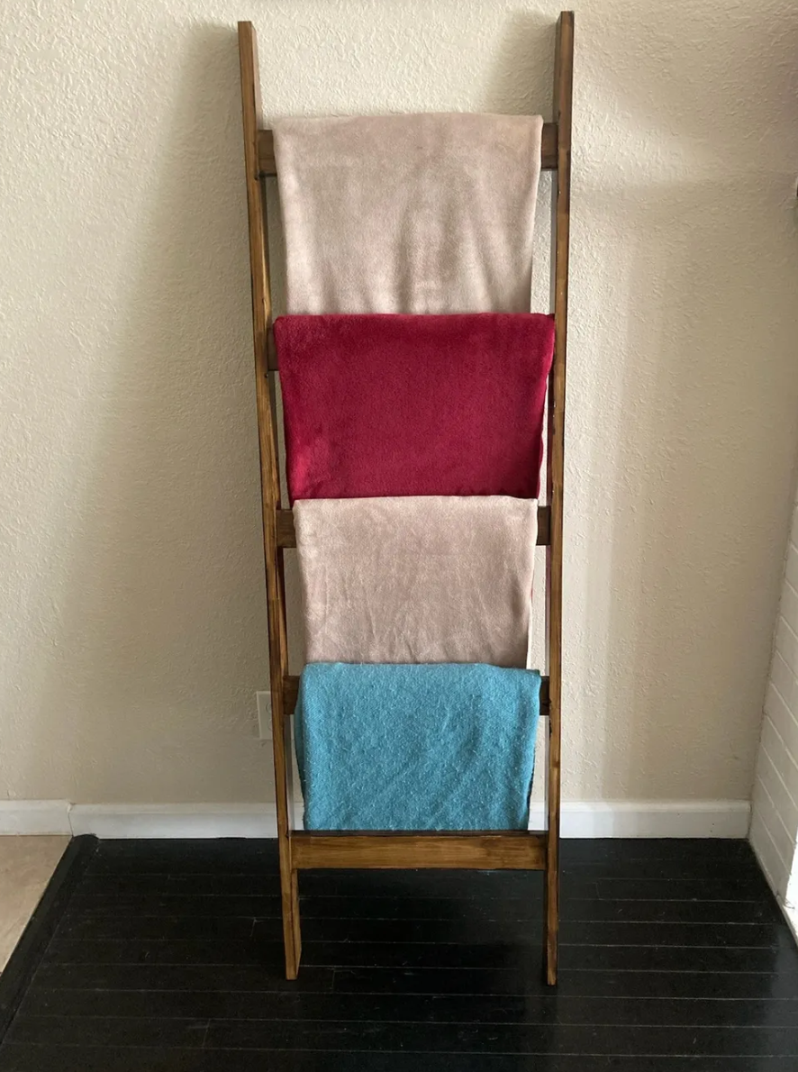 A wooden blanket ladder with four colorful towels hanging on it, standing against a beige wall on a dark floor.