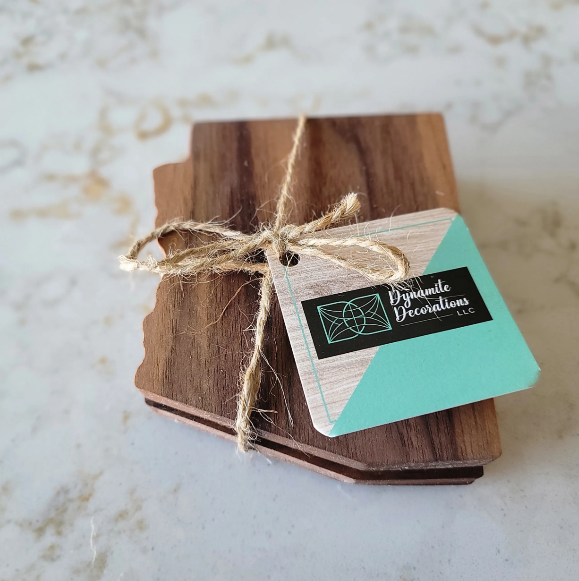 A small wooden sign tied with a twine bow, attached to a business card for 'Dynamite Decorations LLC,' resting on a piece of wood.