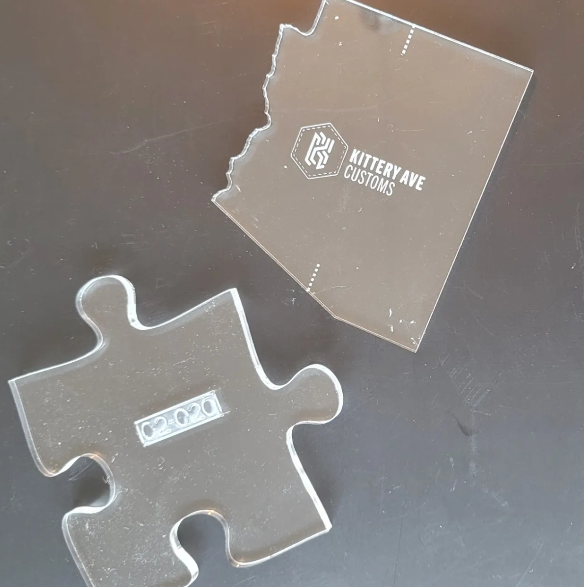 A clear acrylic puzzle piece with the code '0202' and the same piece's detached top section with the logo and text 'Kittery Ave Customs' engraved on it, placed on a dark surface.