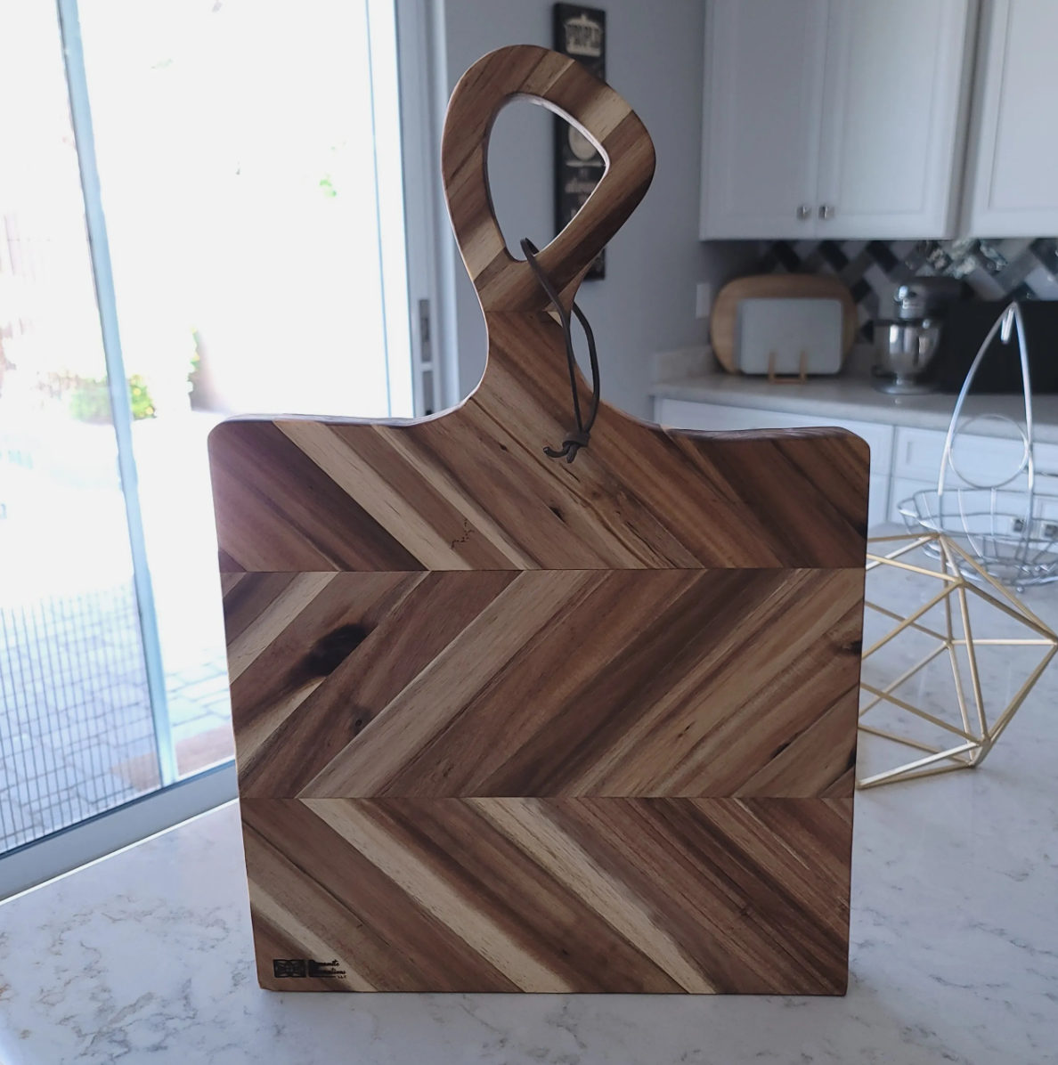 Wooden cutting board with a handle in a kitchen, next to a gold geometric wire basket and a glass jar. Kitchen cabinets and a sliding door are in the background.