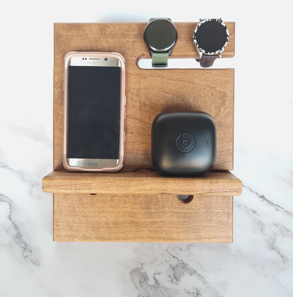 Wooden organizer on marble surface holding a smartphone, two smartwatches, and a wireless earbuds case.