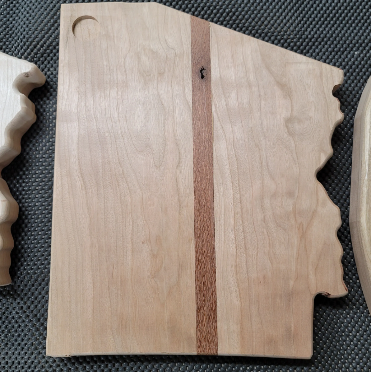 Unfinished wooden cutting board with a central strip of darker wood, placed on a black textured surface.