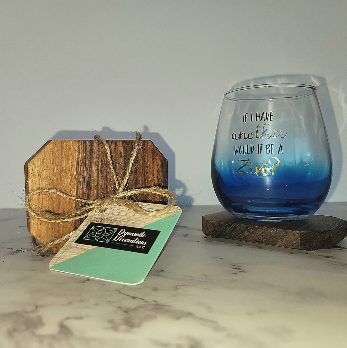 Decorative blue glass with gold and black lettering reading, 'If I have another, would it be a Zinf?,' placed on a small dark wooden coaster, with a wooden block tied with jute string and a business card from Dynamite Decorations LLC on a white marbl